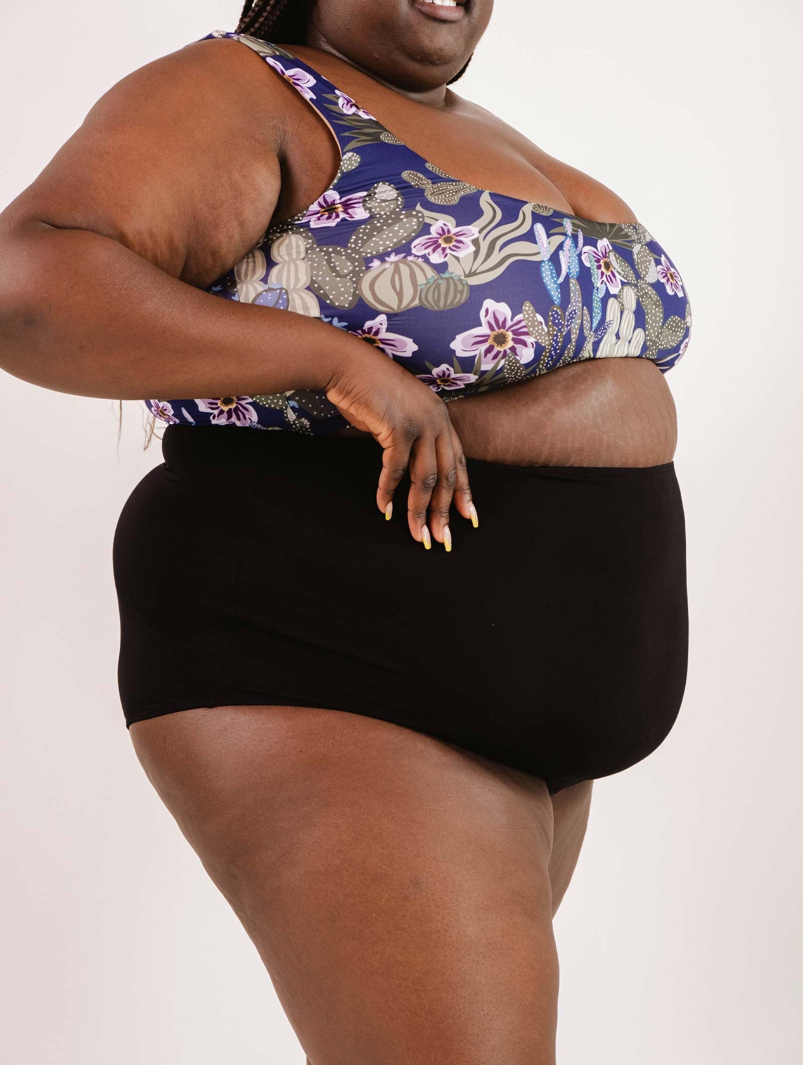 Person wearing a floral sports bra and Bermudes Black extra high waisted bikini bottom by Mimi & August, presenting a flattering silhouette against a white background.