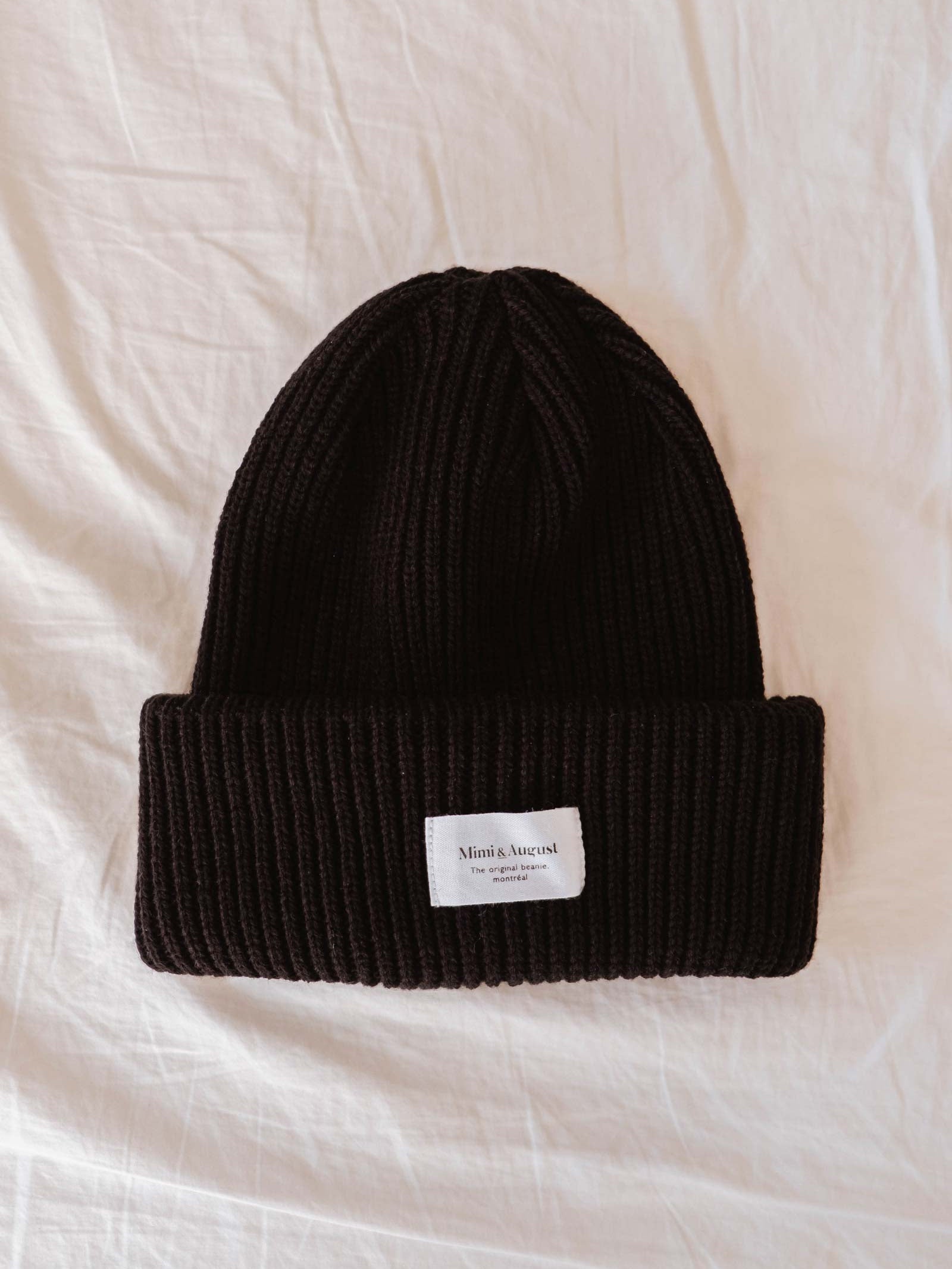 A Black Snuggle Beanie from Mimi & August, featuring a ribbed knit design and a white label on the cuff, lays flat on a white fabric surface, making it the perfect oversized winter accessory.