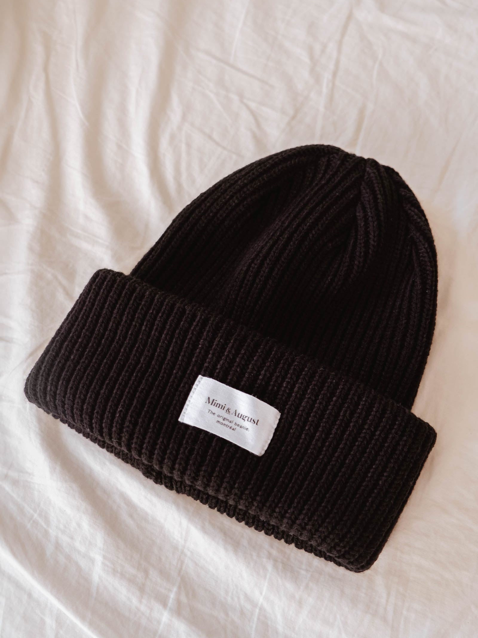 A Black Snuggle Beanie from Mimi & August, adorned with a small white label, lies on a white fabric surface, making it the perfect winter accessory.