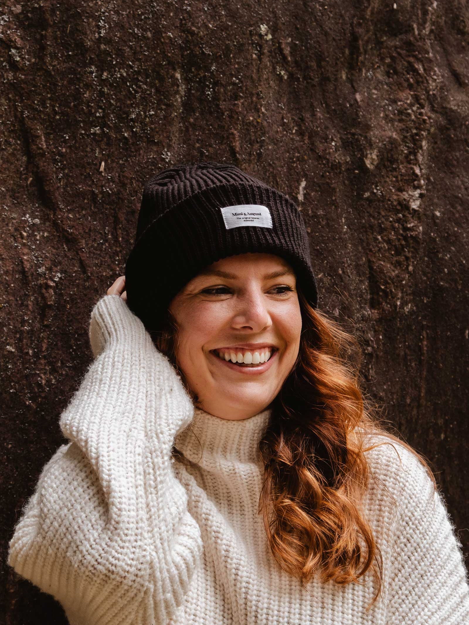 A woman with long hair is smiling while wearing a cream sweater and a cozy Black Snuggle Beanie by Mimi & August, standing in front of a textured wall.
