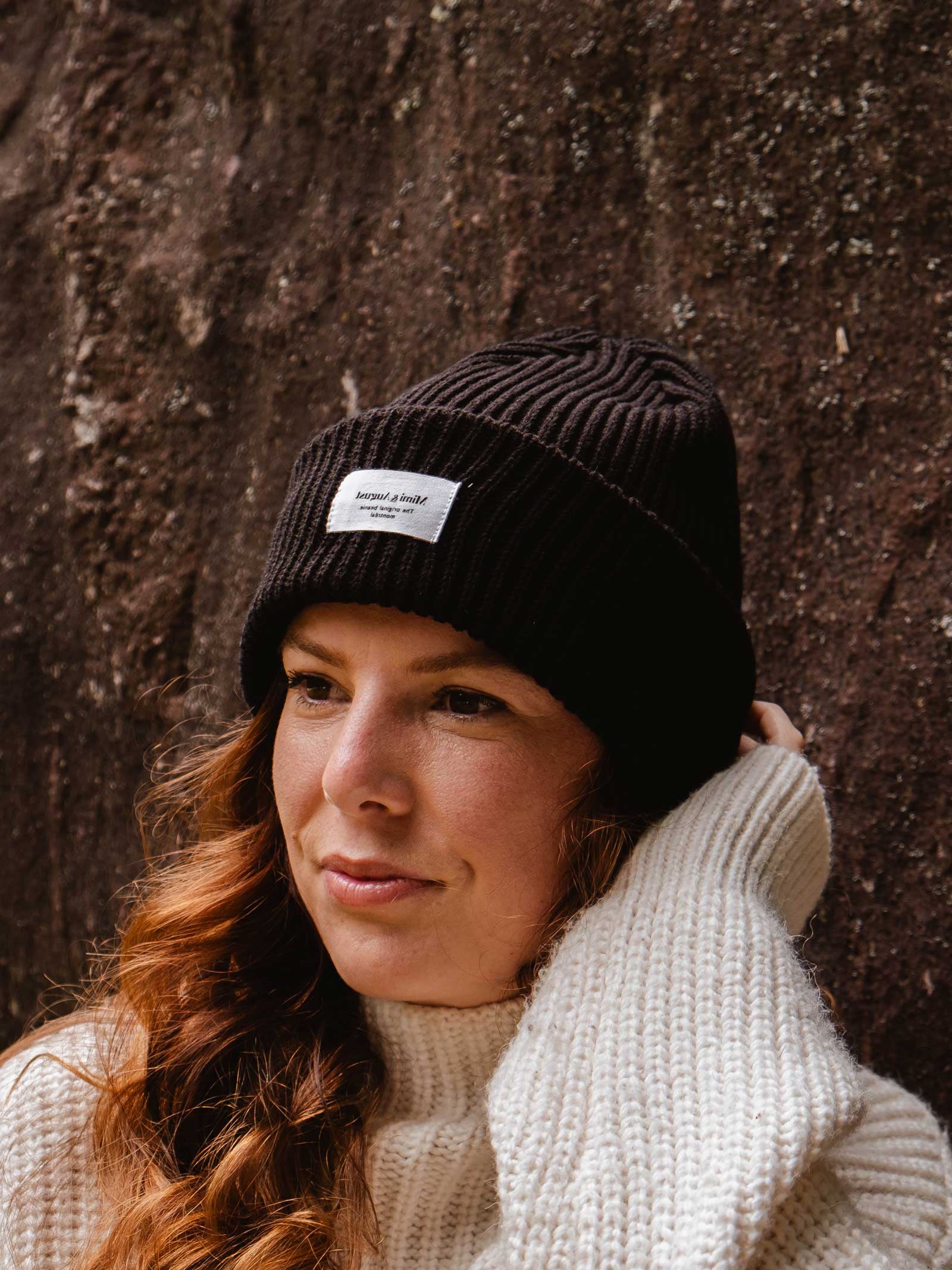 A person with long wavy hair, wearing a Black Snuggle Beanie by Mimi & August and a white sweater, stands against a rough-textured wall.