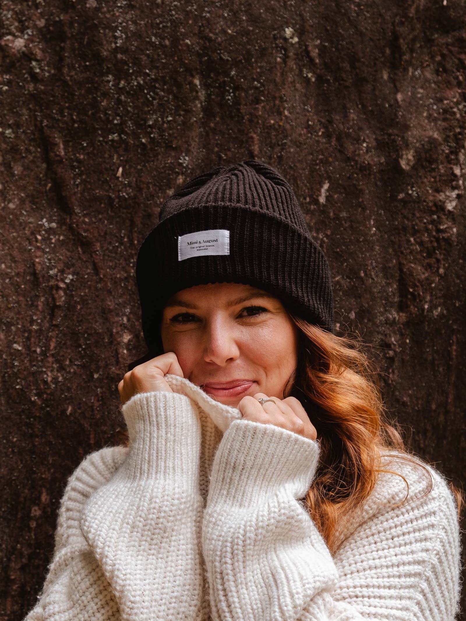 A person with long hair wears a Black Snuggle Beanie from Mimi & August and a white knit sweater, standing in front of a textured dark background, smiling and holding the sweater collar close to their face.