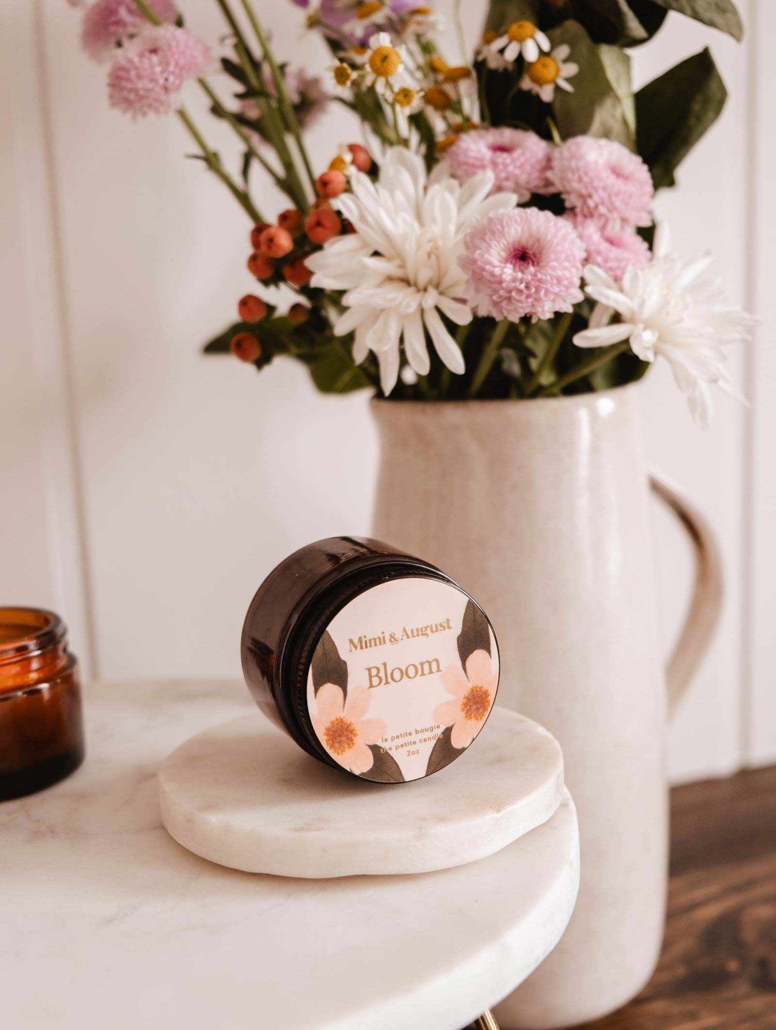 Une bougie de soya réutilisable Bloom de Mimi & August repose sur une surface ronde en marbre, emplissant l'air de son arôme fruité, tandis qu'un vase de fleurs roses et blanches illumine l'arrière-plan.