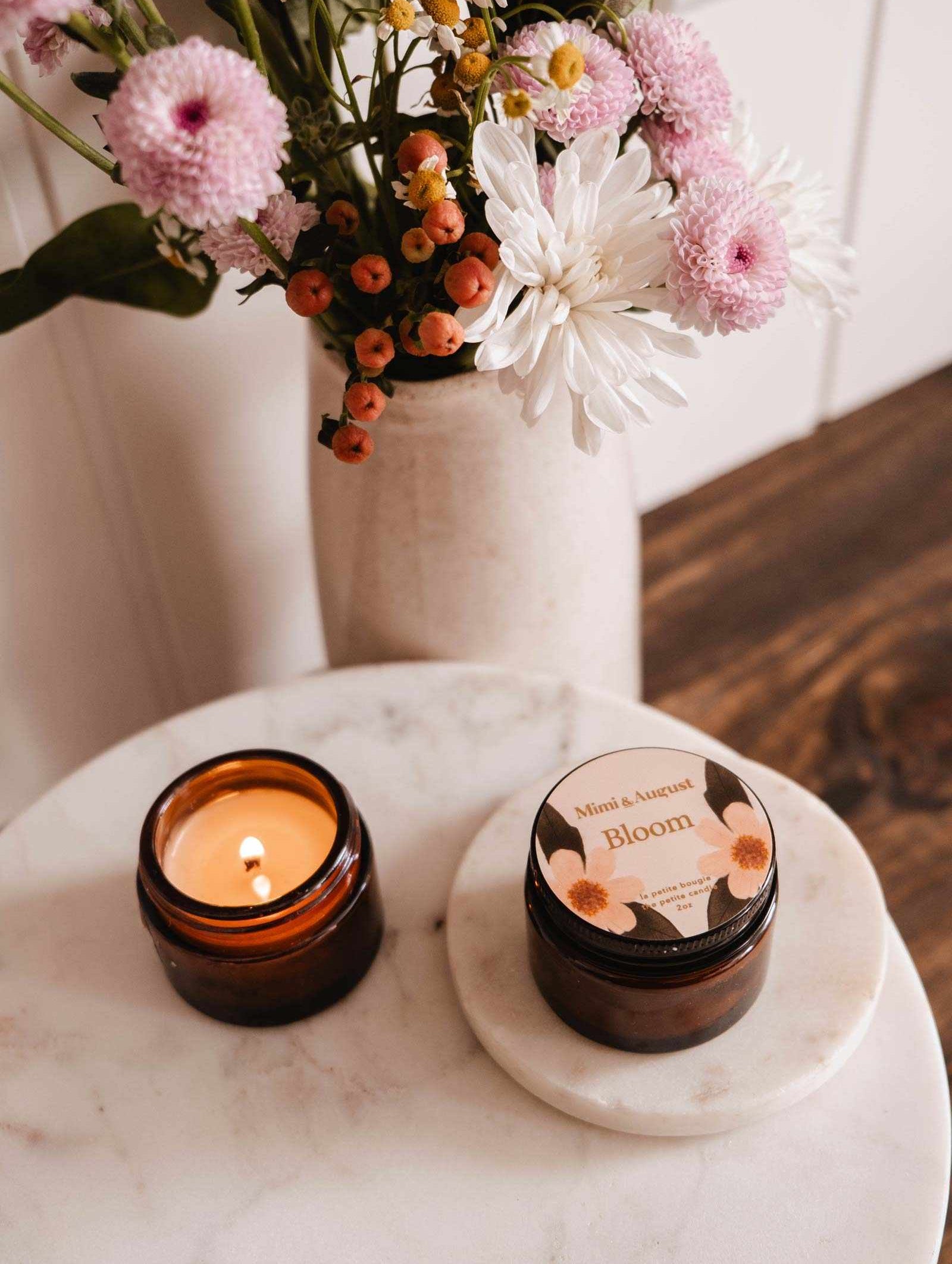 Une bougie de soya réutilisable Bloom de Mimi & August allumée dans un pot ambré repose sur du marbre à côté d'un pot Bloom fermé, avec un vase de fleurs roses et blanches en arrière-plan.