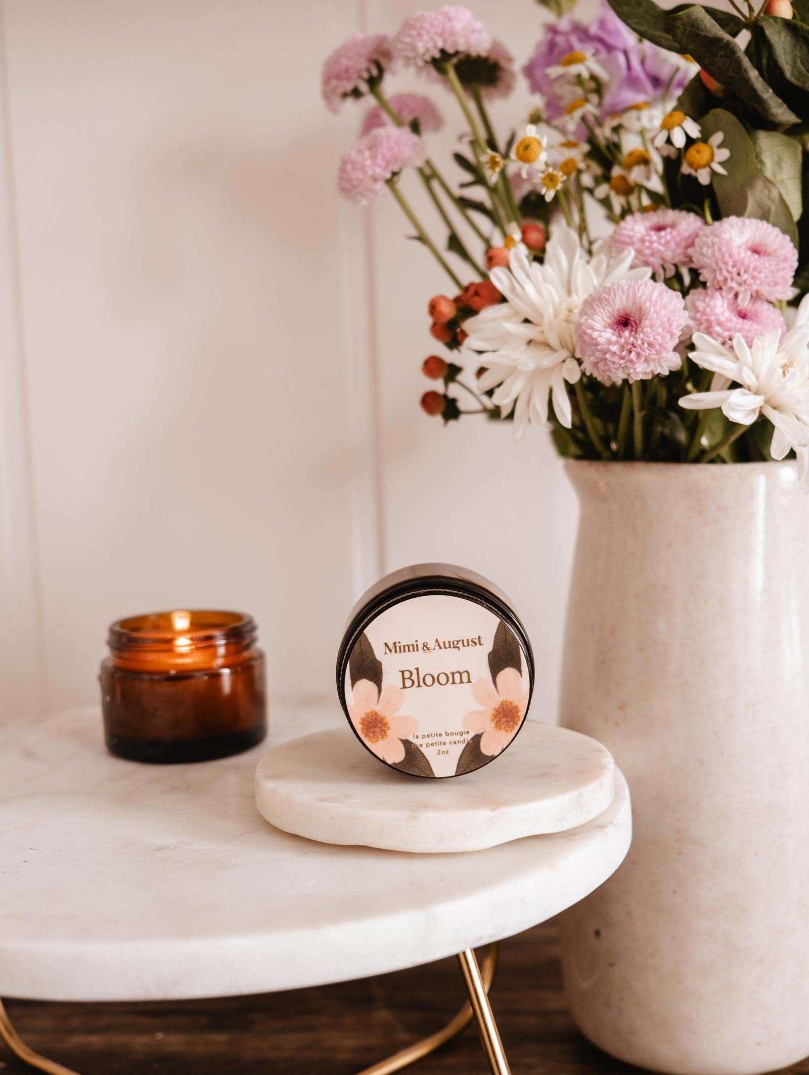 Une bougie de soya réutilisable Bloom de Mimi & August repose sur une surface en marbre blanc à côté d'un pot de bougie brun allumé et d'un vase de fleurs roses et blanches.