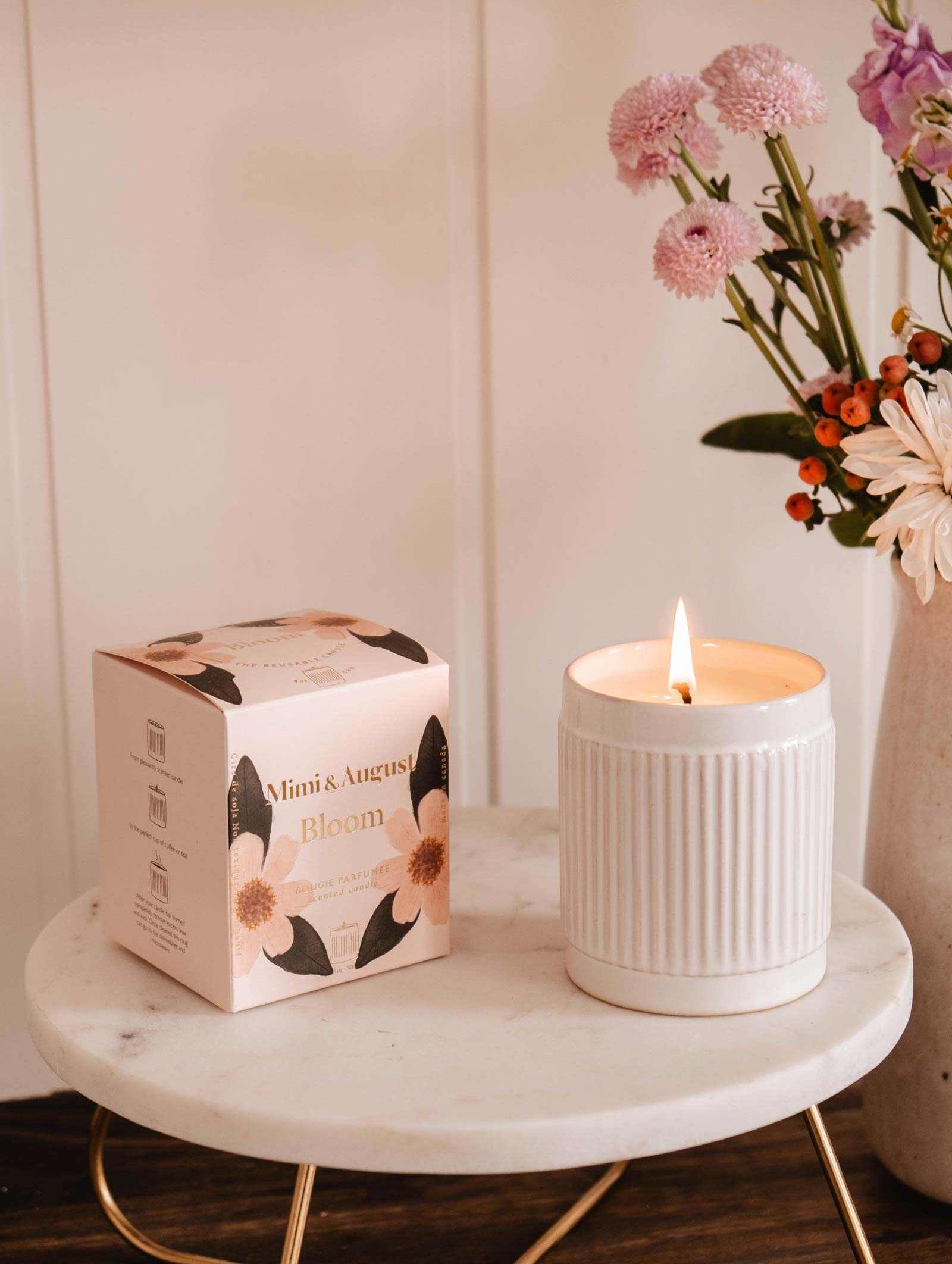 The Mimi & August Bloom – Reusable Soy Candle, orange cranberry scented in a white ceramic holder with floral packaging, is displayed on a round marble table beside a vase of pink and orange flowers.
