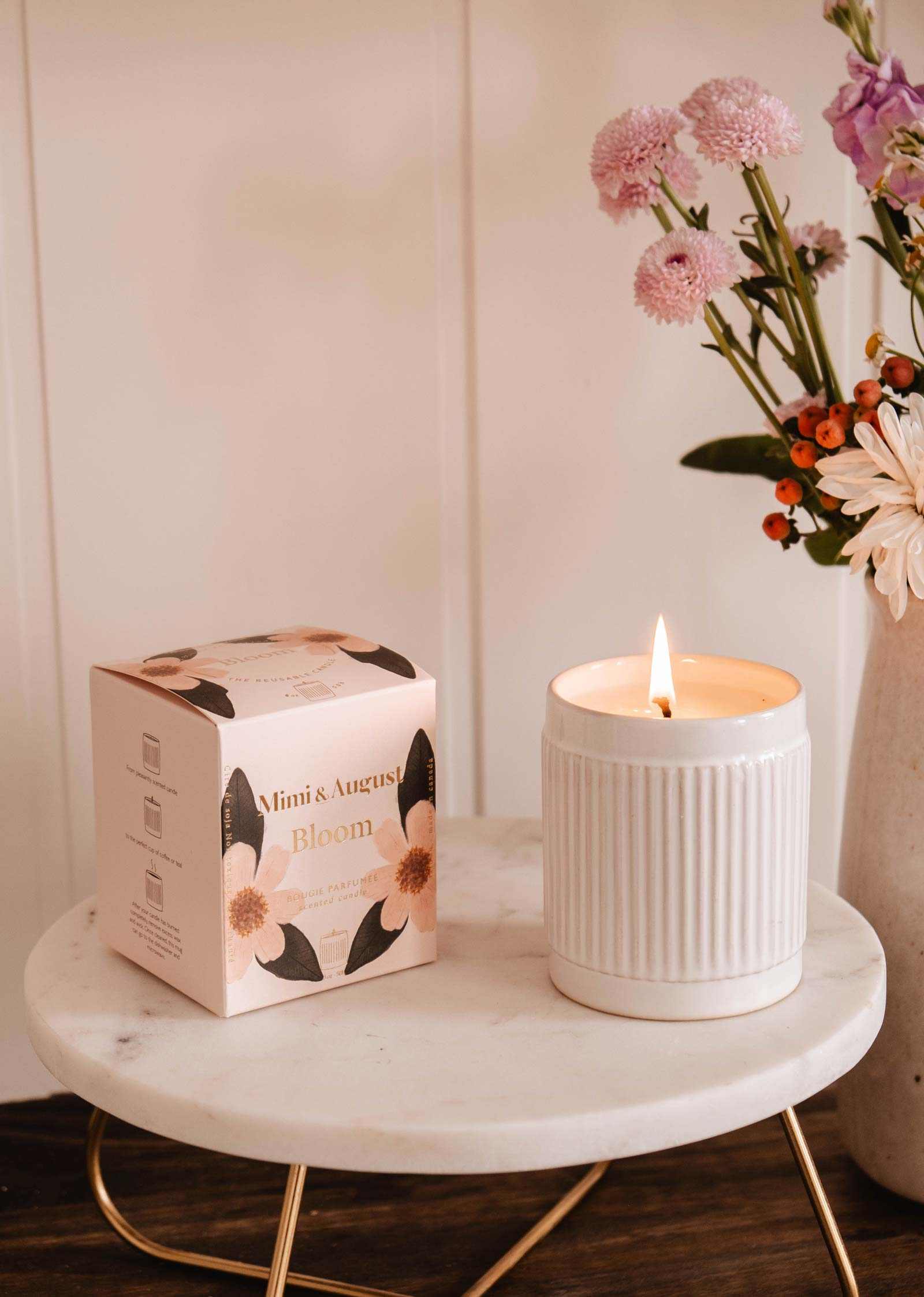 The Mimi & August Bloom – Reusable Soy Candle, orange cranberry scented in a white ceramic holder with floral packaging, is displayed on a round marble table beside a vase of pink and orange flowers.