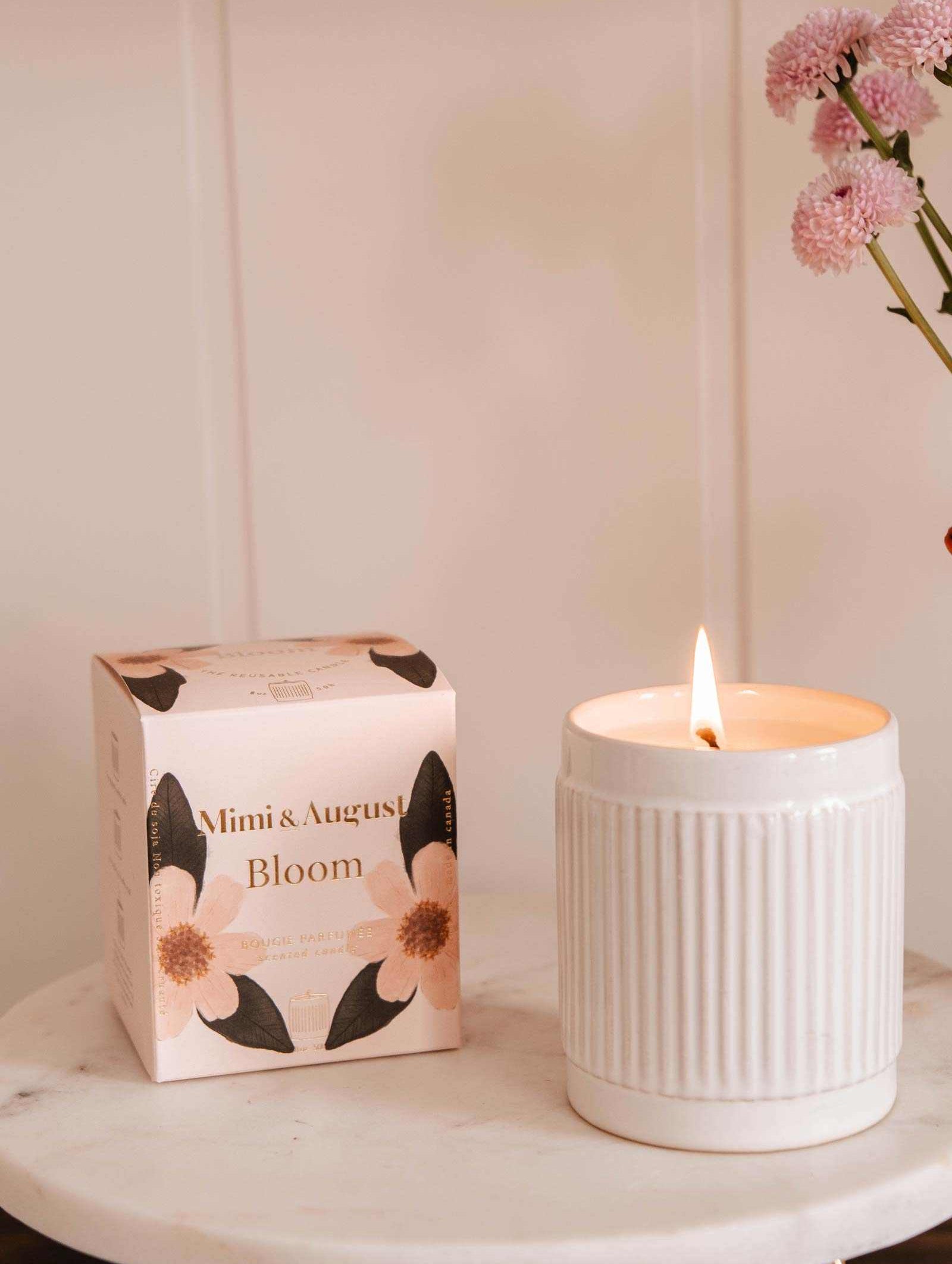 A lit Mimi & August Bloom – Reusable Soy Candle with an orange cranberry scent sits on a marble surface beside a floral "Bloom" box, with pink flowers in a vase in the background.