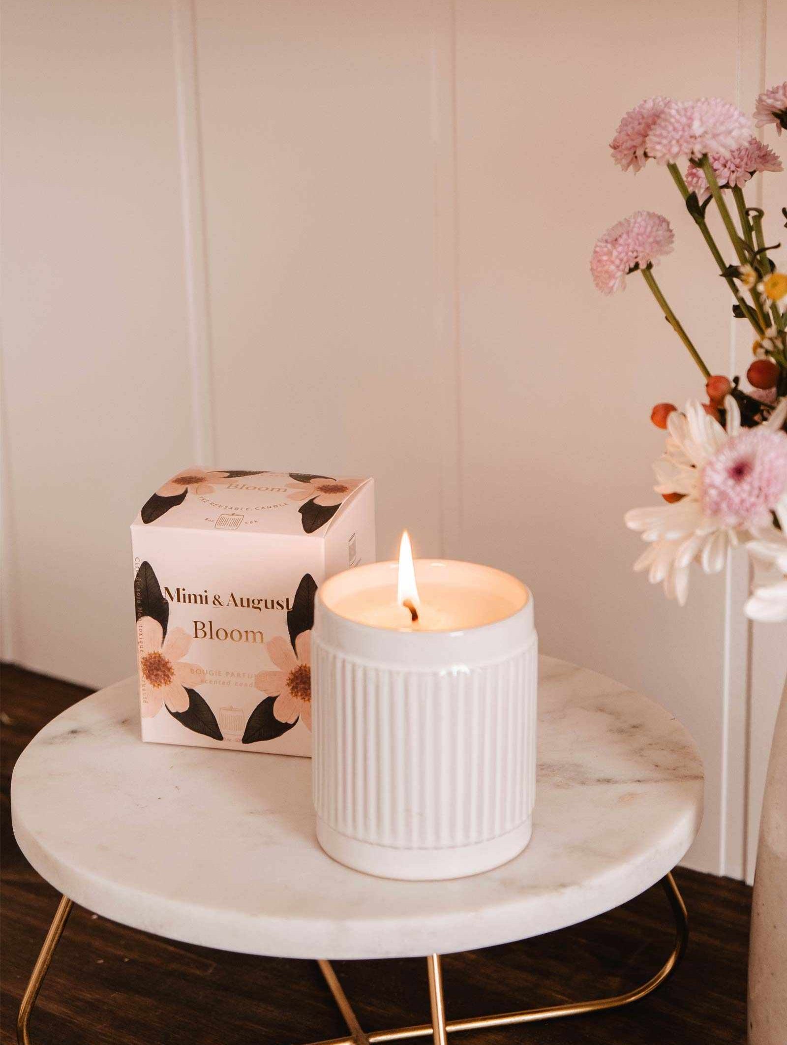 The Bloom – Reusable Soy Candle by Mimi & August, in a ribbed holder, glows on a round marble table beside a floral box and pink flowers in a vase.