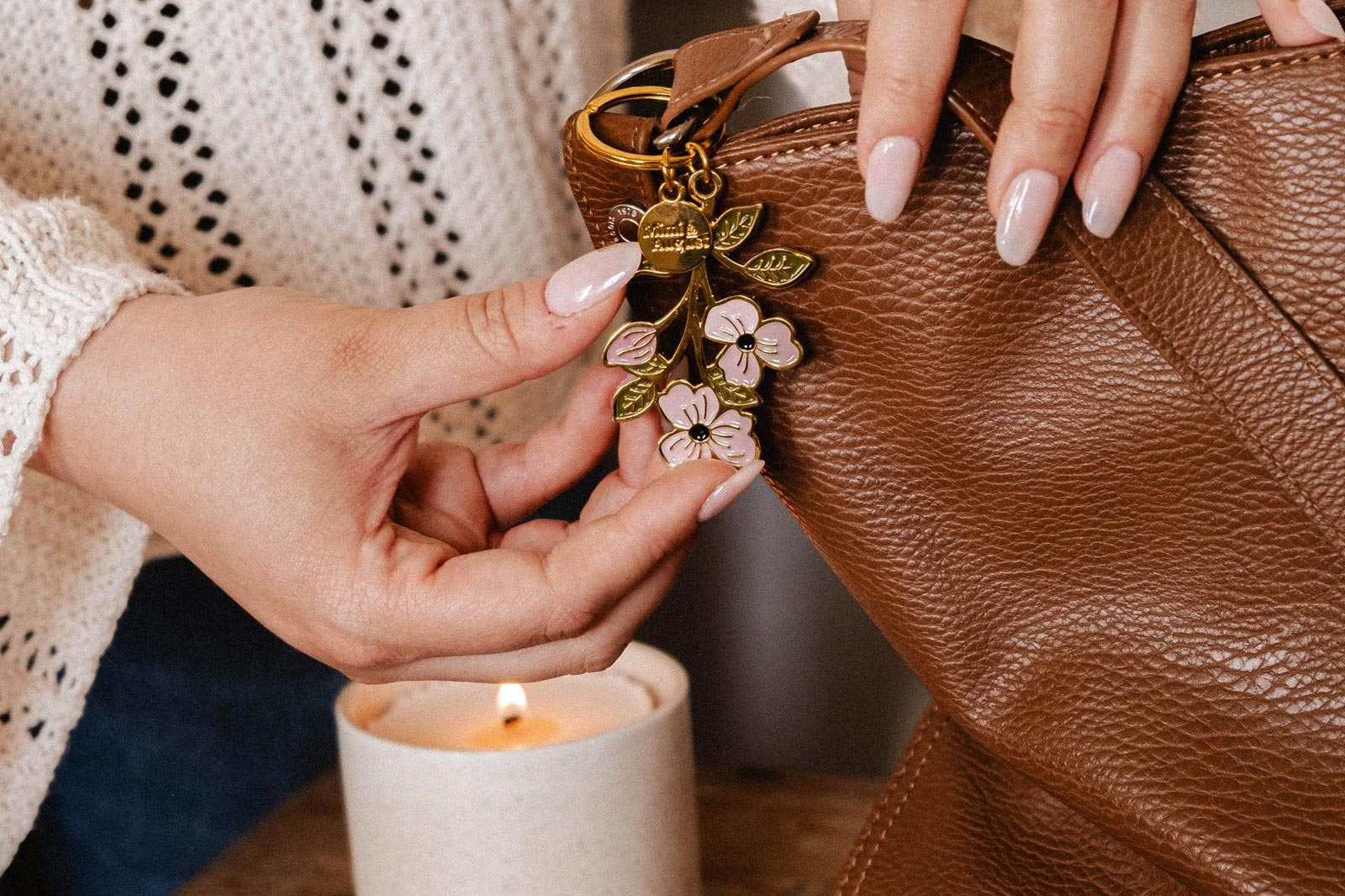 Someone attaches a Mimi & August Blossom Branch Keychain to a brown leather bag placed on a table next to a lit candle, a notebook, and a pen.