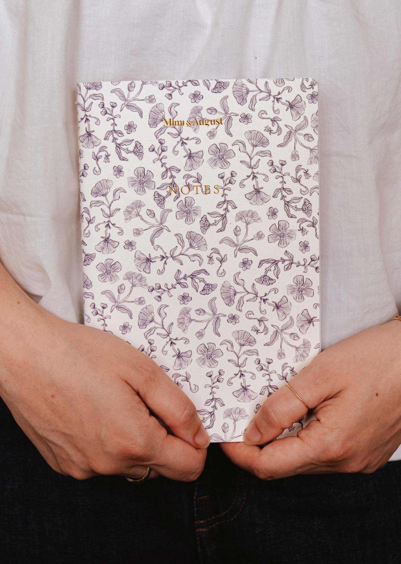Someone holding the Blossom - Notebook by Mimi & August, featuring a purple floral pattern and gold "NOTES" text on the cover.