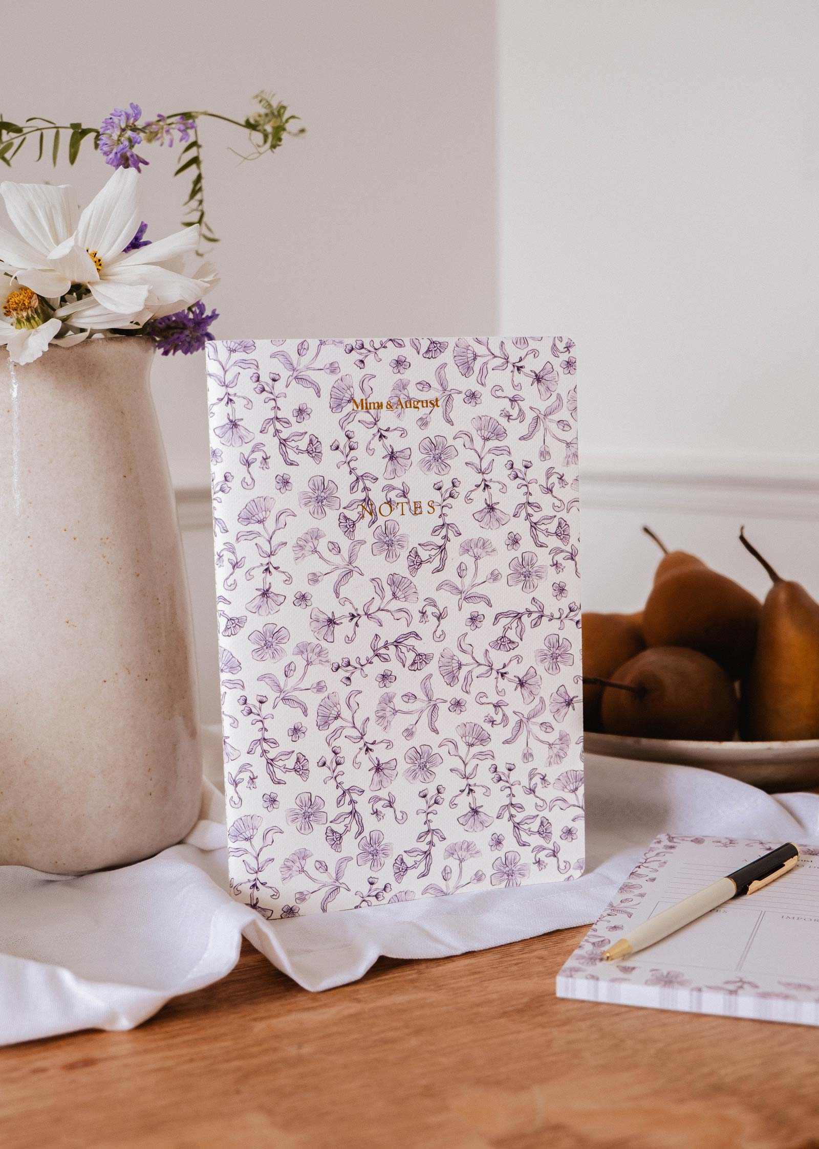 The Blossom - Notebook by Mimi & August, made from recycled paper, stands upright on a wooden table beside a vase of flowers, a plate of pears, and a lined journal with a pen.