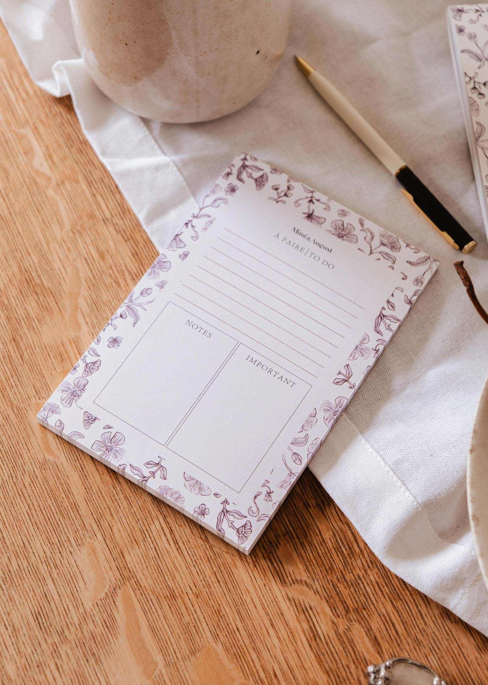 The Blossom - Notepad by Mimi & August, crafted from recycled paper with a floral design, sits on a wooden table next to a pen, white cloth, and ceramic pieces.