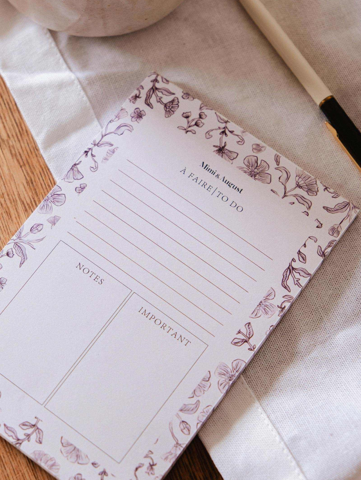 The Blossom - Notepad by Mimi & August, made from recycled paper and featuring sections for notes and important items, is displayed on a cloth surface next to a pen.
