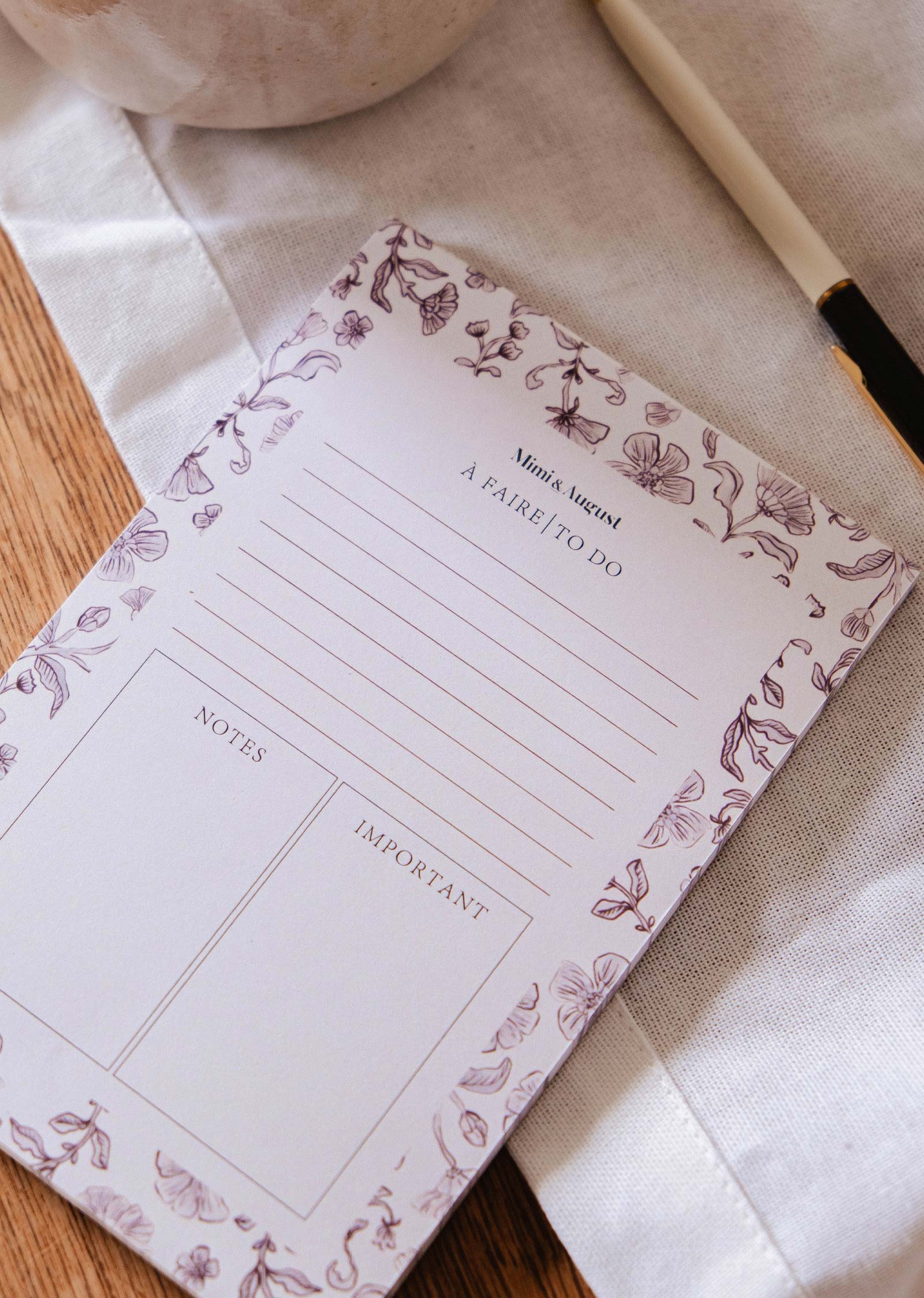 The Blossom - Notepad by Mimi & August, made from recycled paper and featuring sections for notes and important items, is displayed on a cloth surface next to a pen.