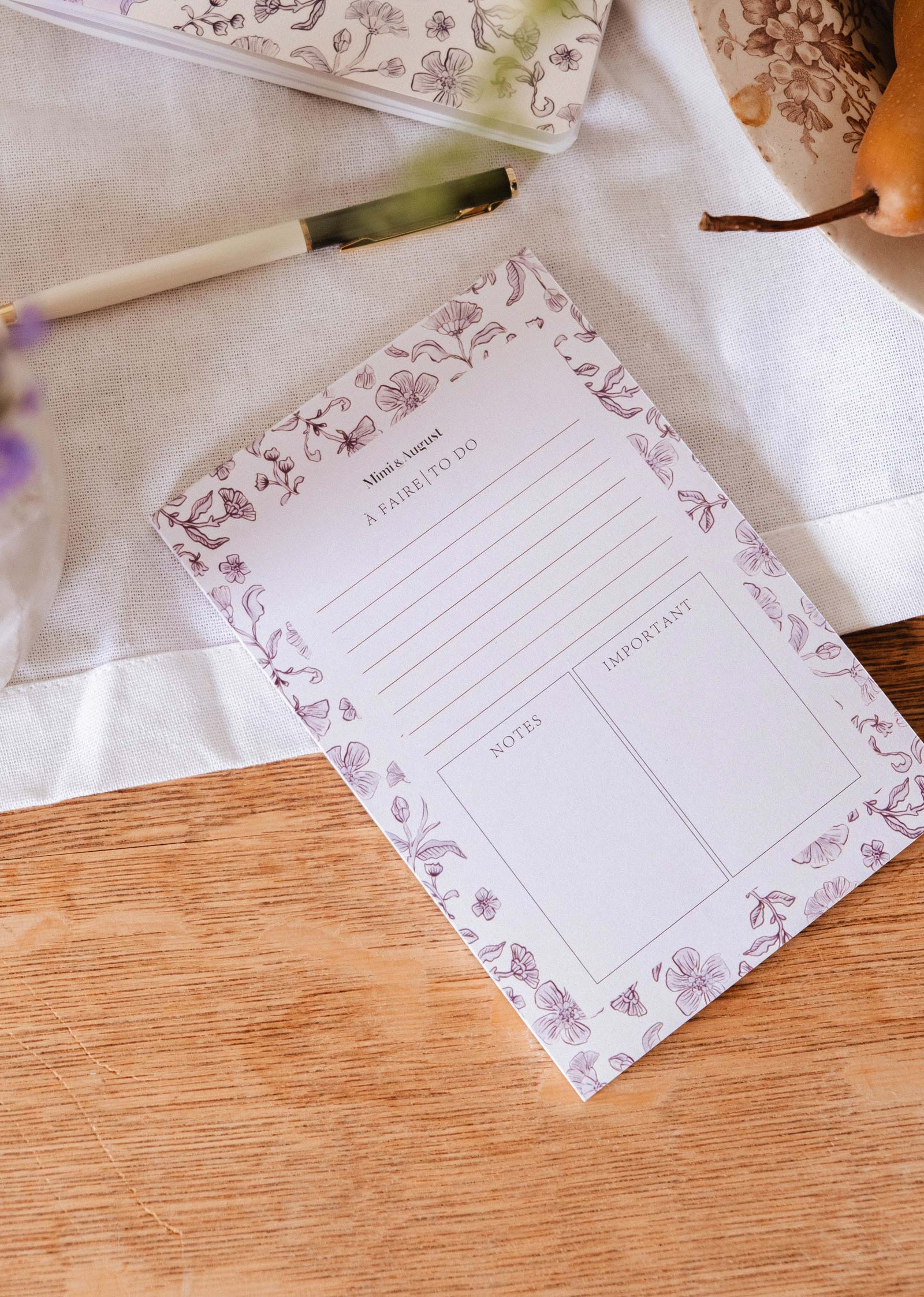 The Blossom - Notepad by Mimi & August, made from recycled paper with sections for to-do lists, notes, and important tasks, rests on a wooden table next to a pen and a plate of fruit.