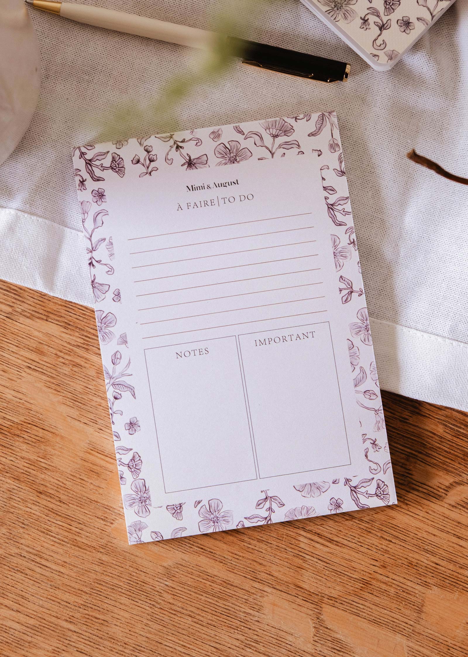 The Blossom - Notepad by Mimi & August, crafted from recycled paper with sections for tasks, notes, and important items, rests on a wooden table beside a pen and a white cloth.