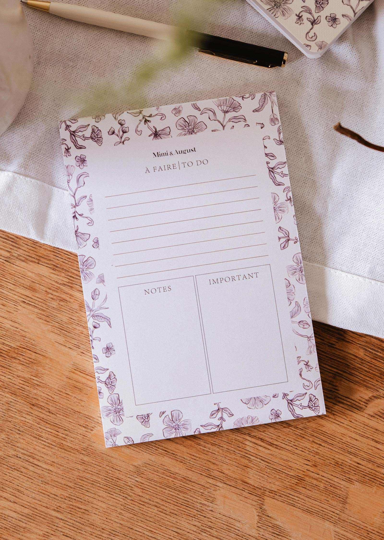 The Blossom - Notepad by Mimi & August, crafted from recycled paper with sections for tasks, notes, and important items, rests on a wooden table beside a pen and a white cloth.