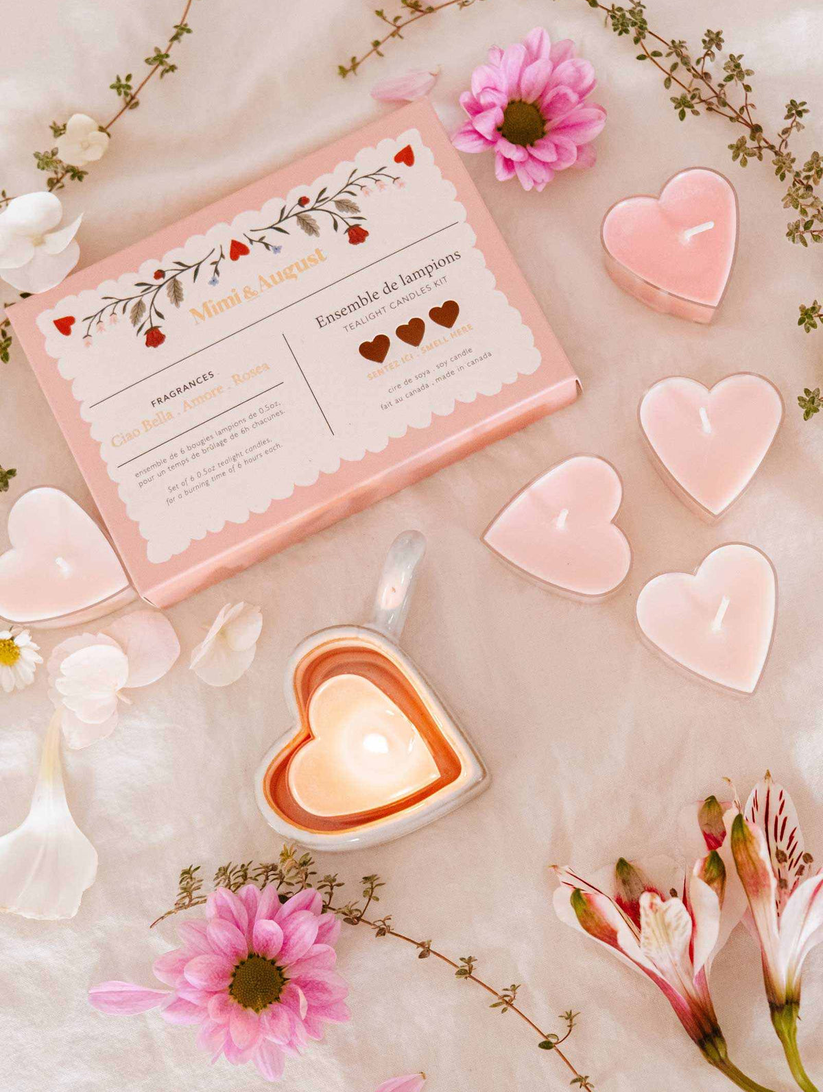 The Mimi & August "Little Hearts – Tealight Candle Set" features heart-shaped soy candles, a lit candle in a heart dish, pink flowers, and white petals elegantly displayed for romantic home decor.