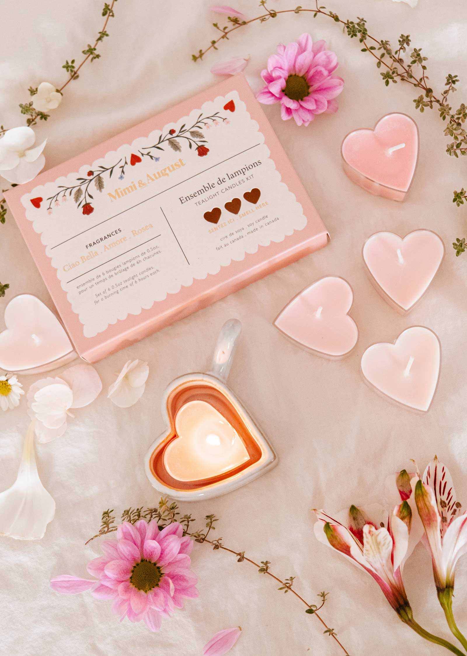 The Mimi & August "Little Hearts – Tealight Candle Set" features heart-shaped soy candles, a lit candle in a heart dish, pink flowers, and white petals elegantly displayed for romantic home decor.