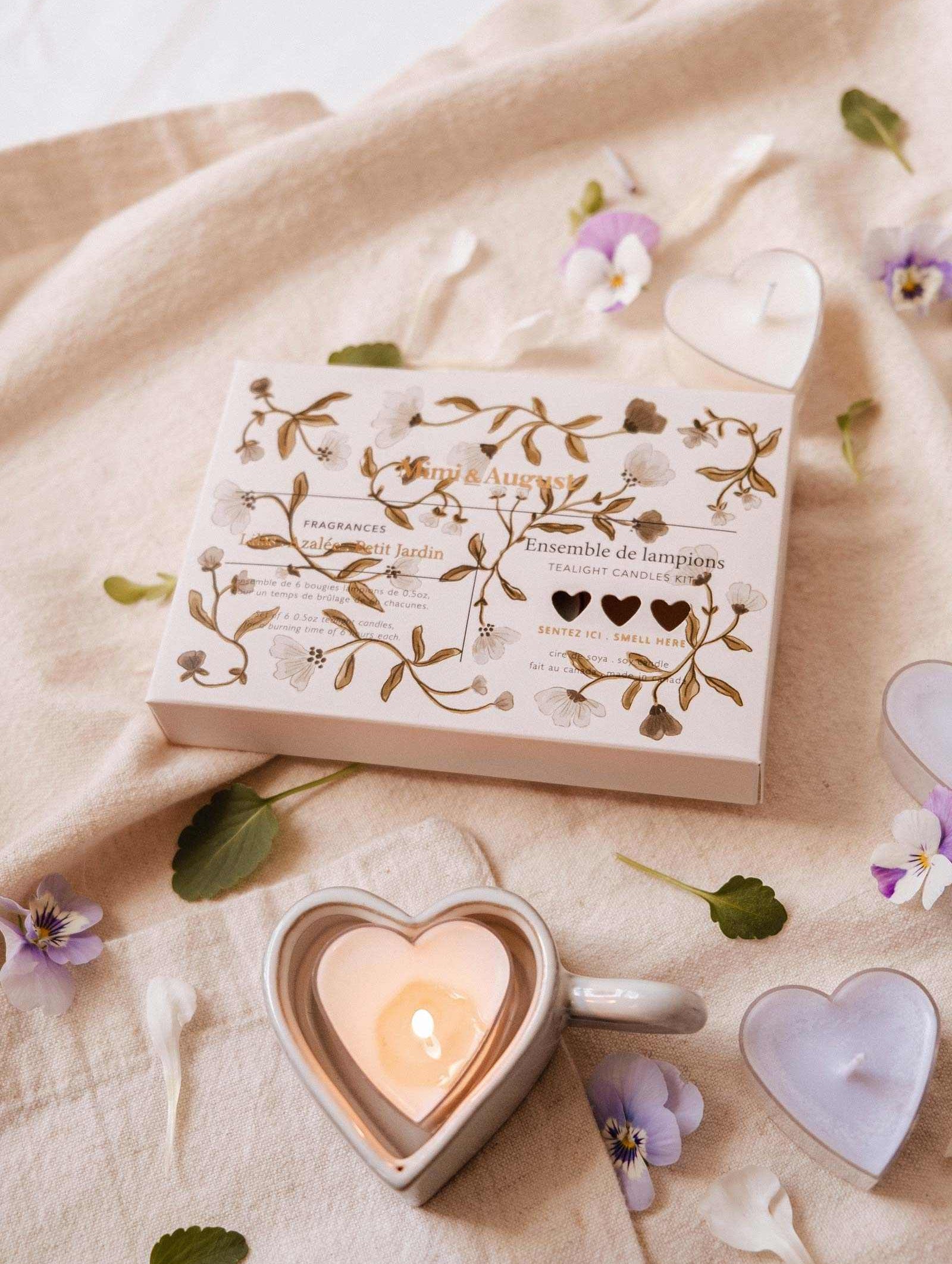 A heart-shaped soy candle from the Mimi & August Blush Hearts Tealight Candle Kit sits in a mug on beige fabric, near its decorative set box and scattered flower petals.