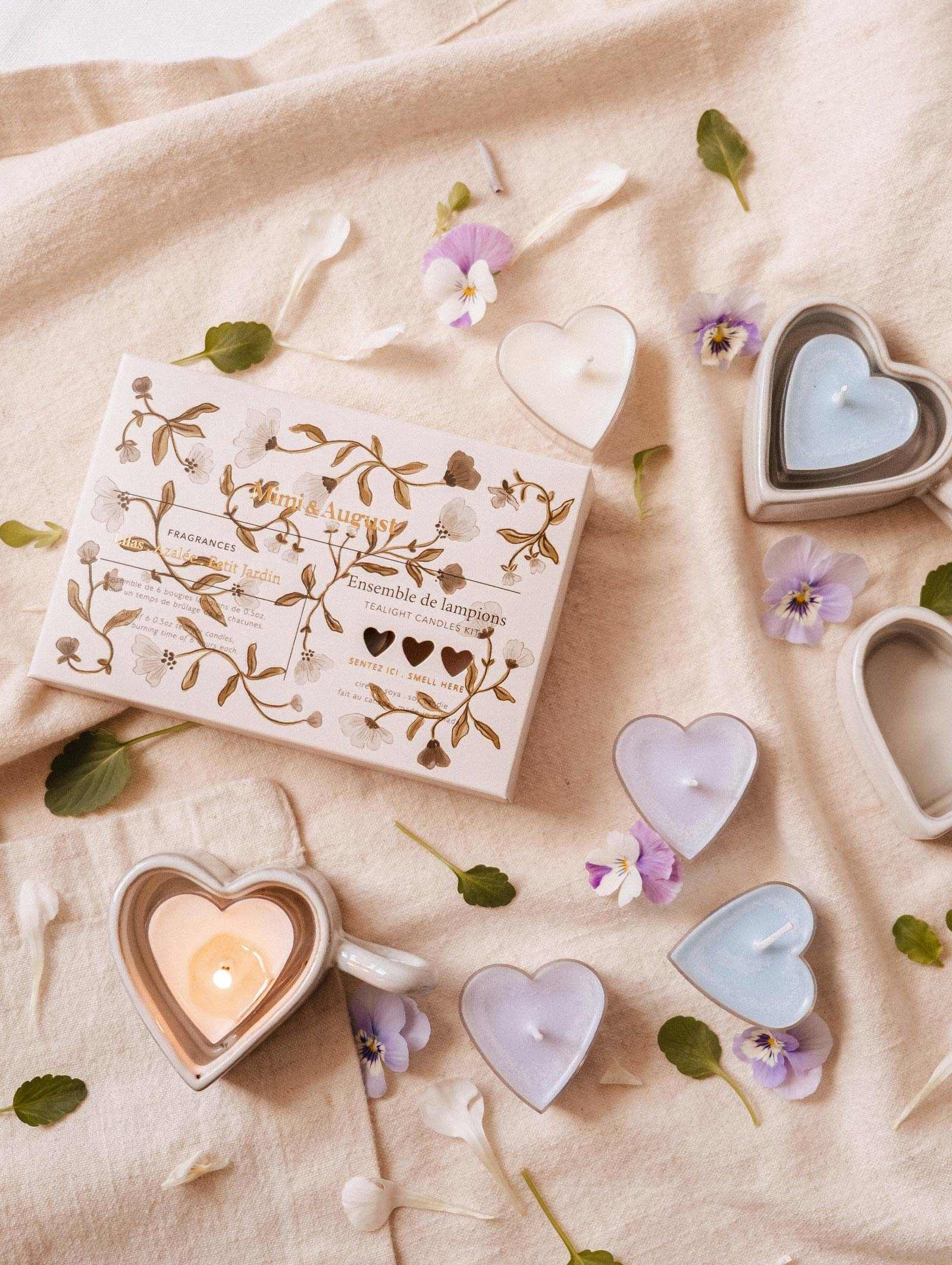 A “Blush Hearts Tealight Candle Kit” by Mimi & August rests on beige cloth, surrounded by hand-poured heart-shaped soy candles in pastel colors and scattered flower petals.