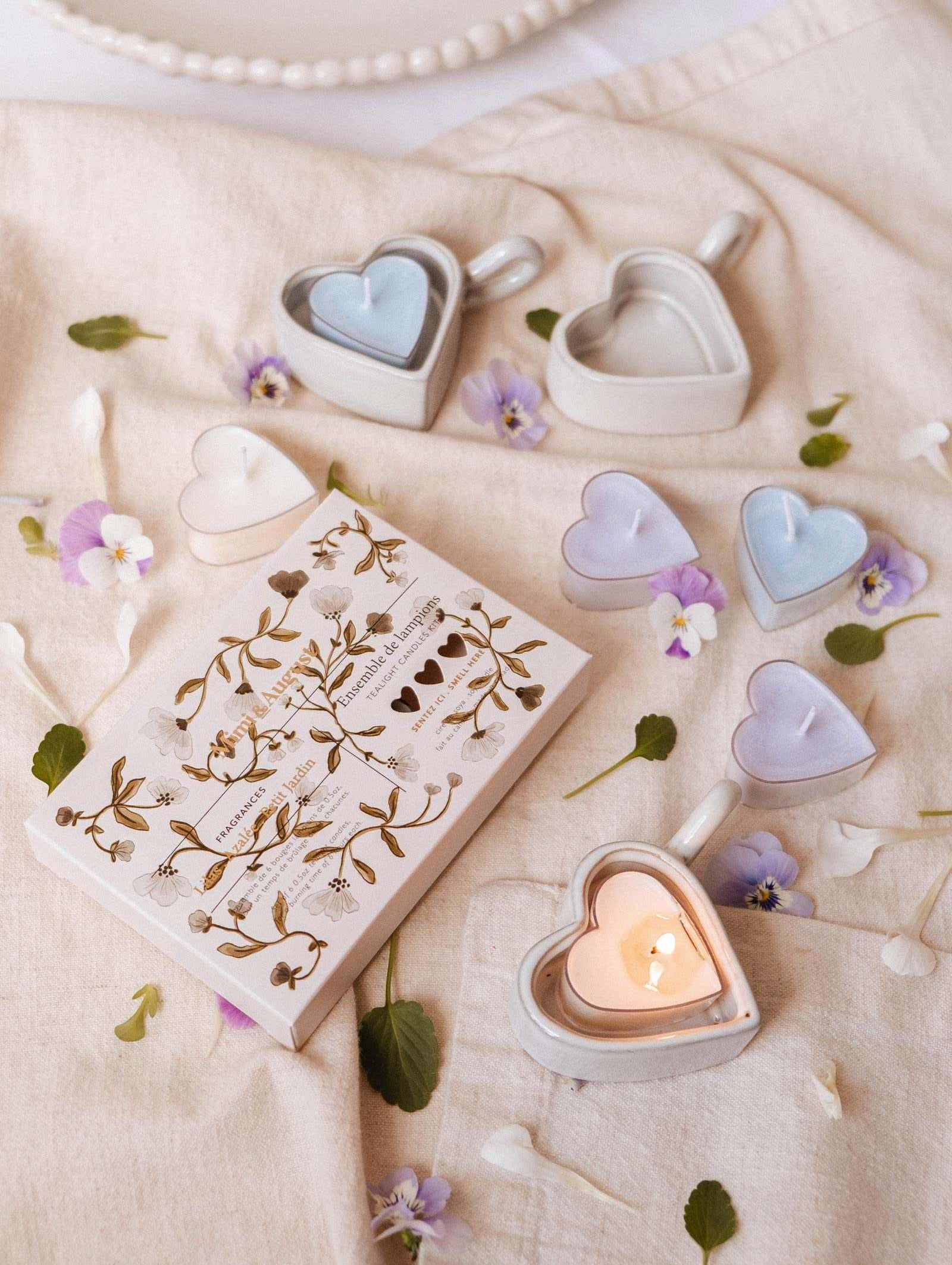 The Blush Hearts Tealight Candle Kit by Mimi & August, featuring heart-shaped soy candles and holders, a decorative book, and scattered flowers and leaves, creates a charming arrangement on light fabric. Hand-poured in Montreal.