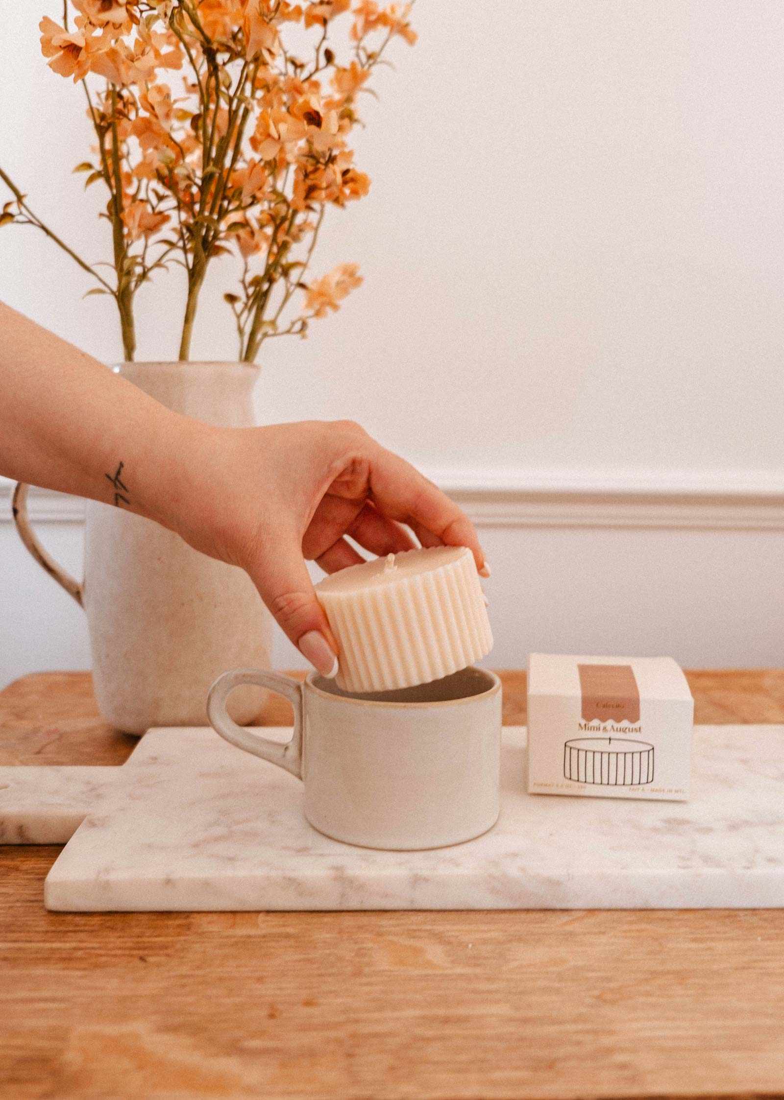 Une main dépose une Recharge de bougie Cafecito cannelée de Mimi & August dans une tasse beige sur une planche en marbre, avec sa boîte et un vase de fleurs orangées en arrière-plan.
