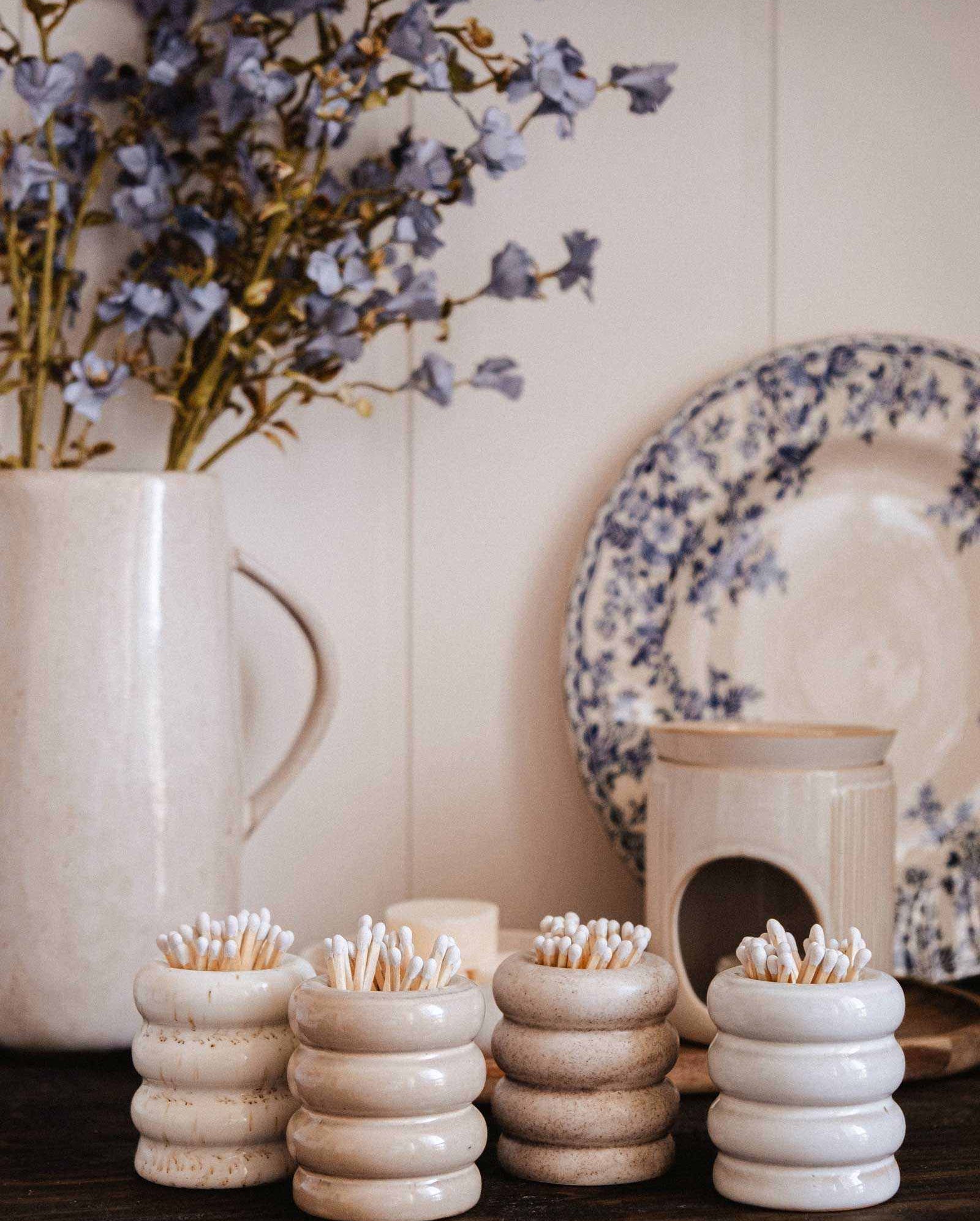 Mimi & August's Ceramic Match Pot holds matches and sits beside cotton-filled ceramic containers, a cream jug with blue flowers, and a floral plate on a wooden surface for a charming display.