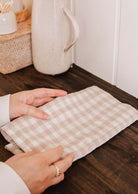 Hands folding the Mimi & August Tea Towel Soft Linen, featuring a beige and white checkered design on a wooden countertop—ideal for drying dishes and keeping your kitchen fresh.