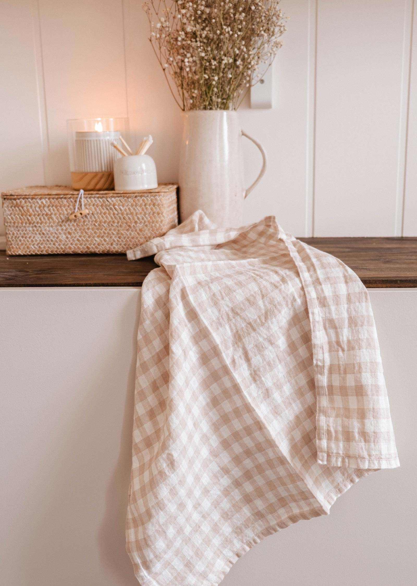 The Mimi & August Tea Towel Soft Linen, in a beige and white checkered pattern, lies on a wooden surface amid dried flowers, a lit candle, and a wicker basket—ideal for drying dishes with its breathable fabric.