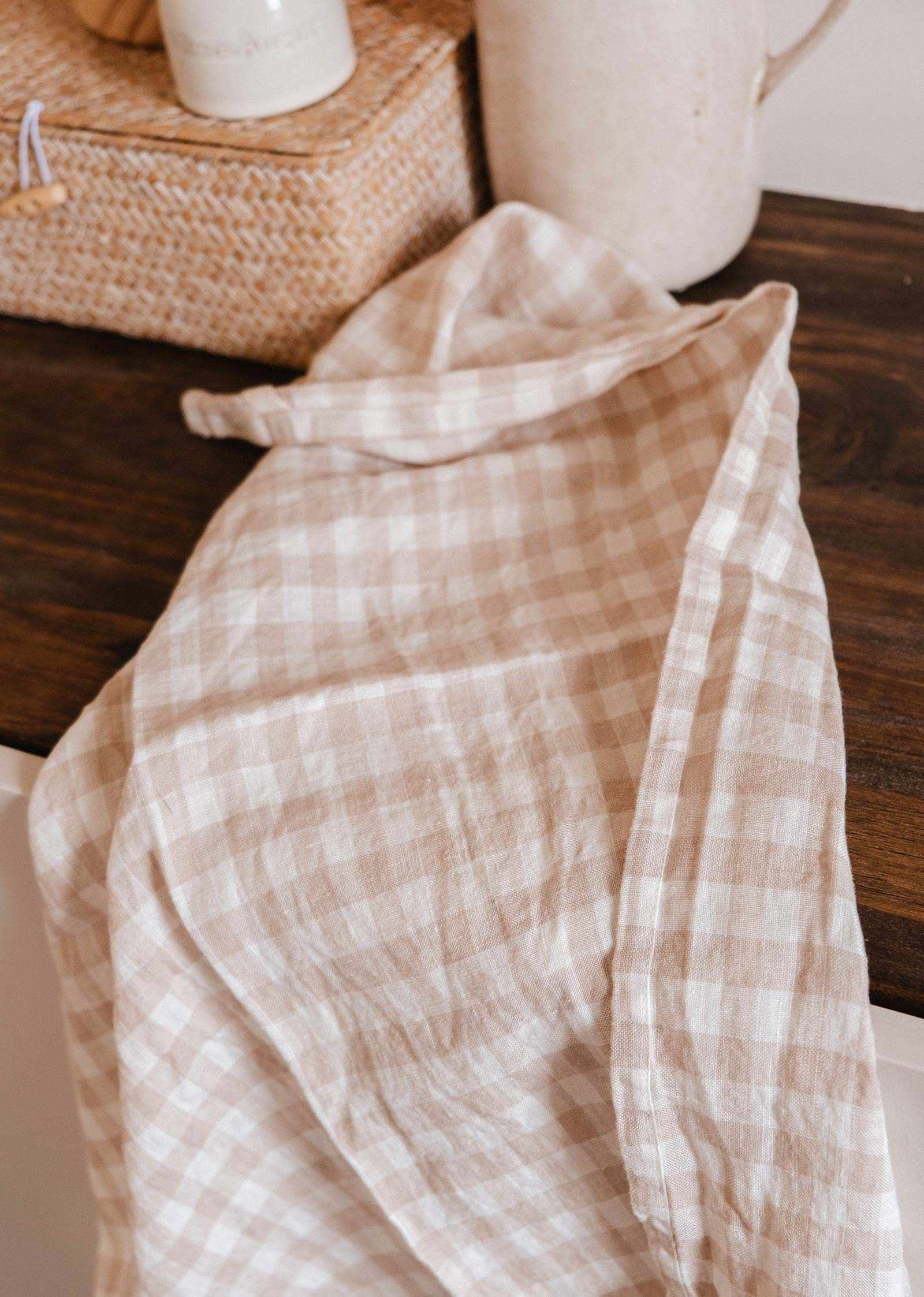 The Mimi & August Tea Towel Soft Linen in beige and white checkered pattern is spread on a dark wooden surface beside a woven basket and a ceramic container.