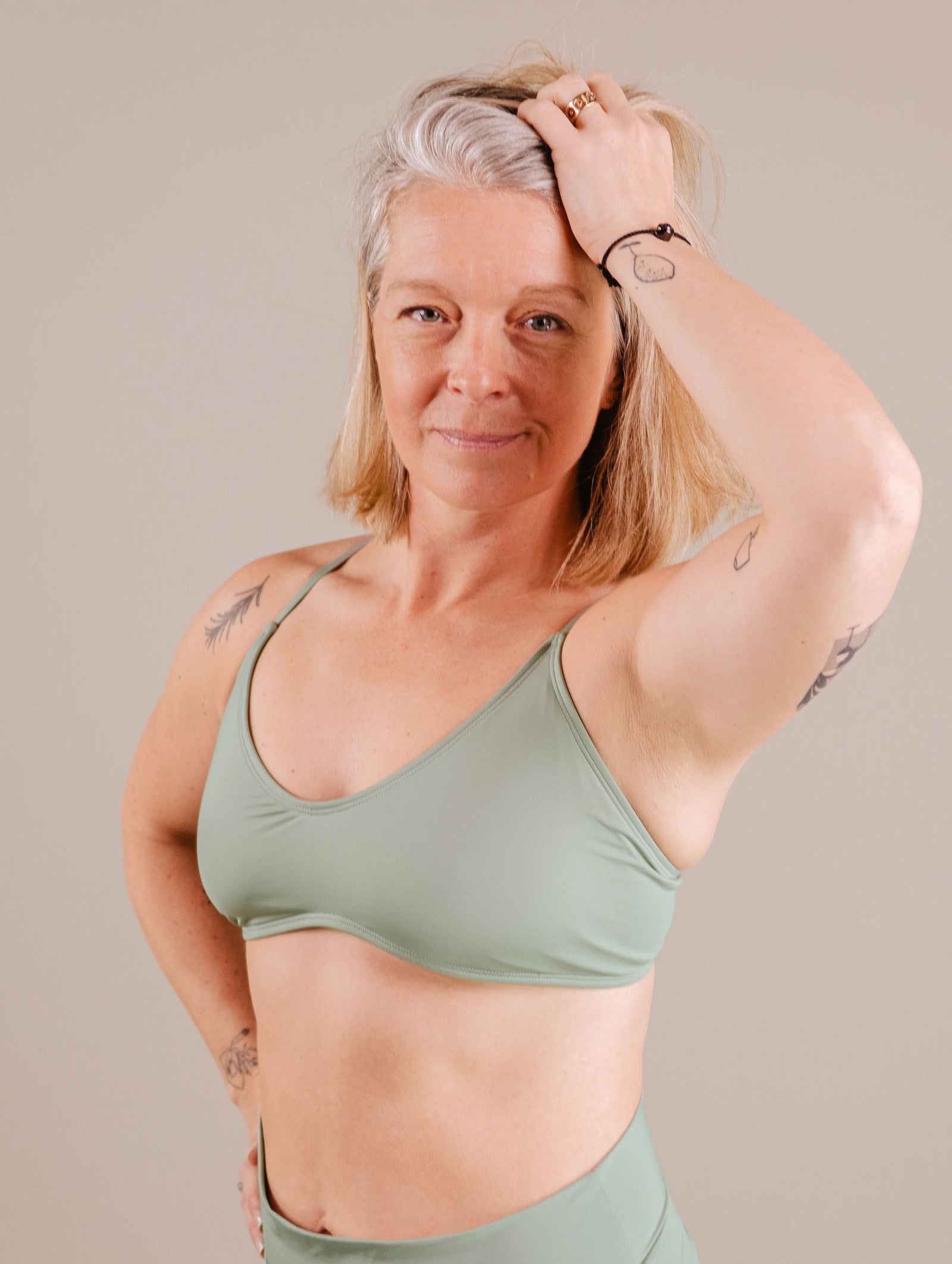 A confident middle-aged woman with gray hair and tattoos, wearing a green Chichi Agave Bralette Bikini Top from Mimi & August and pants, poses with one hand on her head against a beige background.