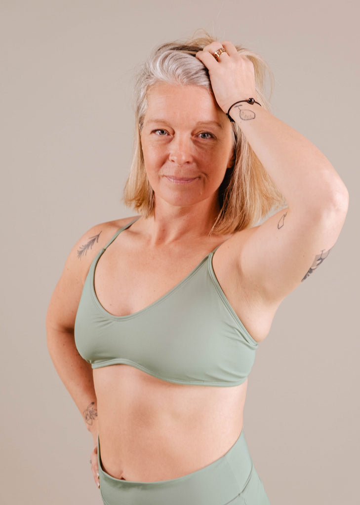 A confident middle-aged woman with gray hair and tattoos, wearing a green Chichi Agave Bralette Bikini Top from Mimi & August and pants, poses with one hand on her head against a beige background.