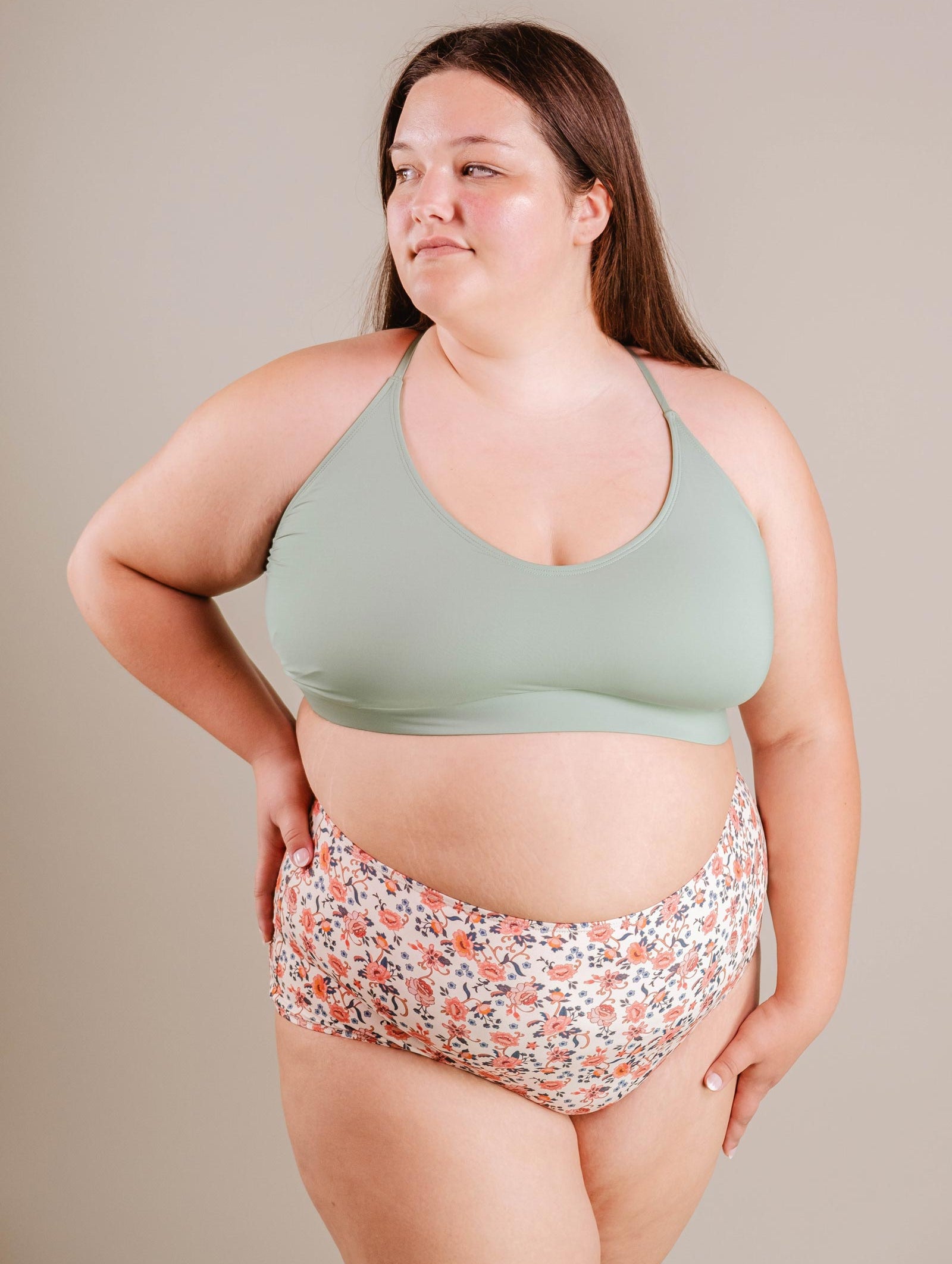 A young woman in a green Mimi & August Chichi Agave Bralette Bikini Top and floral print bikini bottoms stands confidently, looking to the side against a neutral backdrop.
