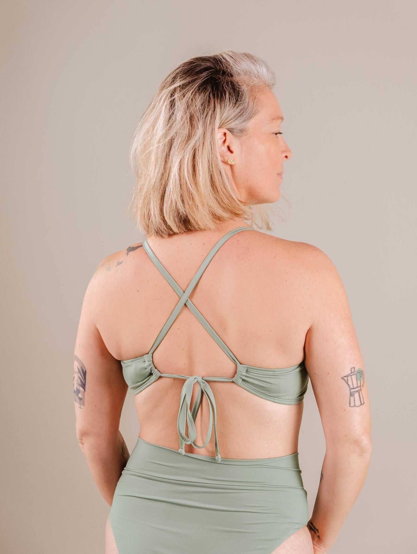 A woman with short blonde hair wearing a green Mimi & August Chichi Agave Bralette Bikini Top swimsuit with a crisscross back, viewed from the side, against a neutral background.