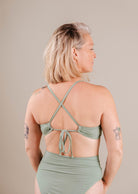 A woman with short blonde hair wearing a green Mimi & August Chichi Agave Bralette Bikini Top swimsuit with a crisscross back, viewed from the side, against a neutral background.