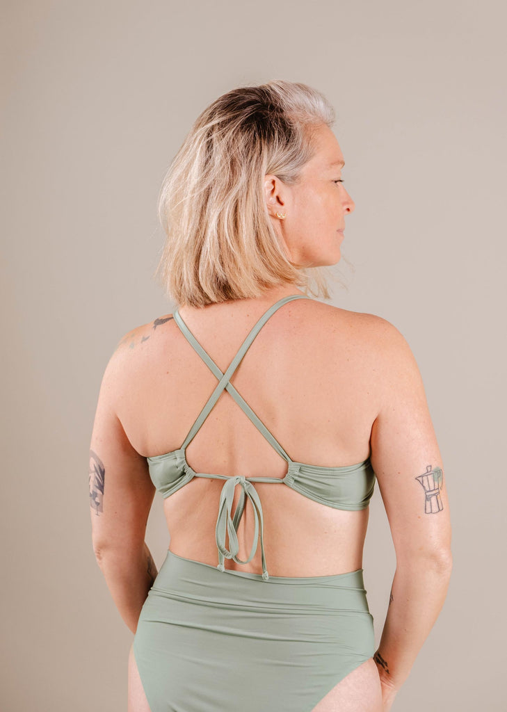 A woman with short blonde hair wearing a green Mimi & August Chichi Agave Bralette Bikini Top swimsuit with a crisscross back, viewed from the side, against a neutral background.