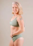 A middle-aged woman with gray hair, wearing a green Chichi Agave Bralette Bikini Top and matching pants, smiling at the camera, showcasing a tattoo on her upper arm by Mimi & August.
