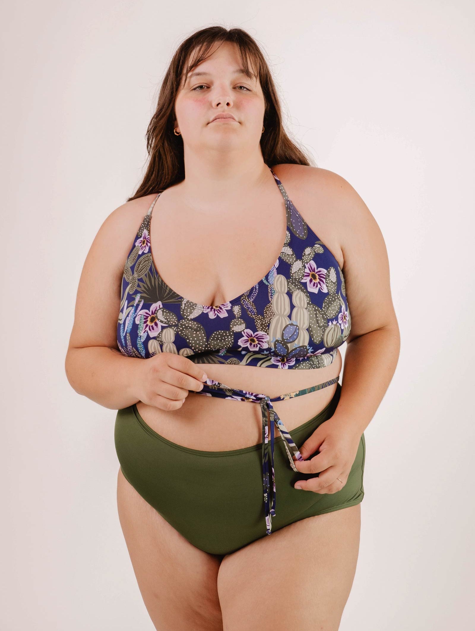 A woman in a green and floral tropical print Mimi & August Chichi Jardin de Nuit - Bralette Bikini Top stands against a plain background, holding the strap of her bikini top as she looks at the camera.