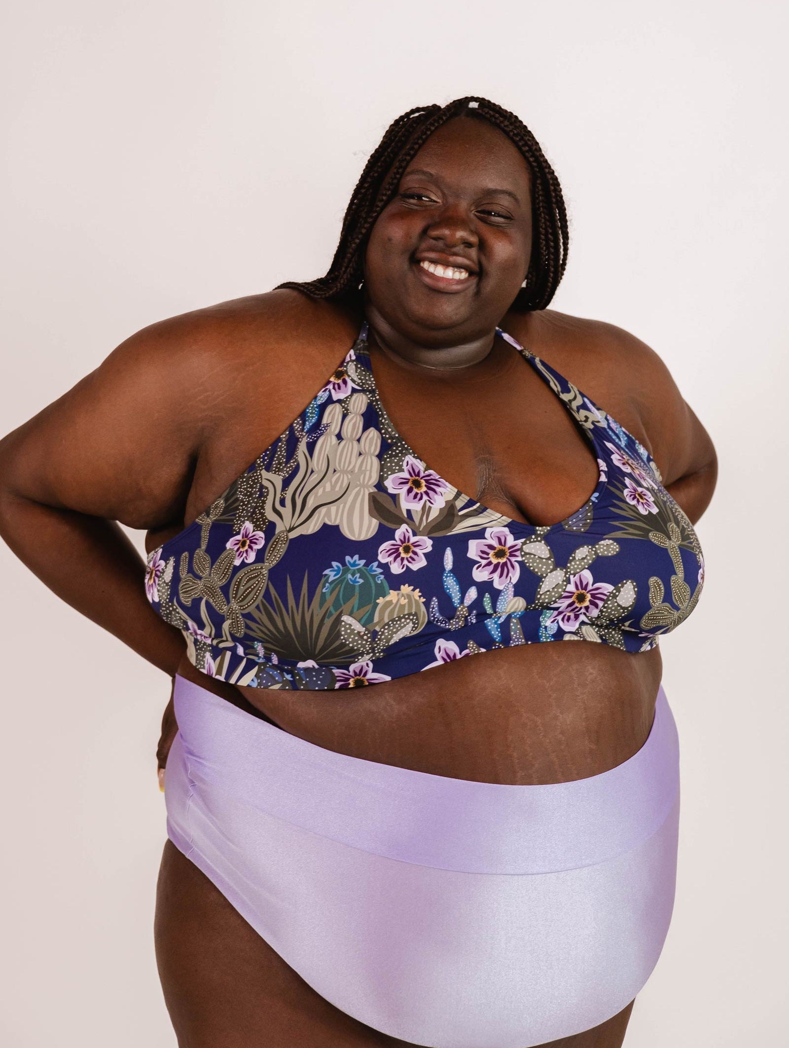 A person with braided hair smiles while wearing a Chichi Jardin de Nuit - Bralette Bikini Top by Mimi & August and light purple bottoms against a plain background.