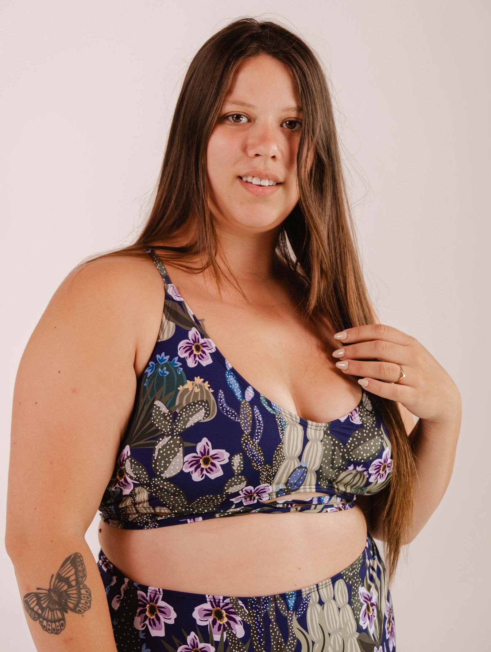 A woman with long brown hair is wearing a Mimi & August Chichi Jardin de Nuit - Bralette Bikini Top with adjustable straps. She has a butterfly tattoo on her arm and is standing against a plain background, looking at the camera.