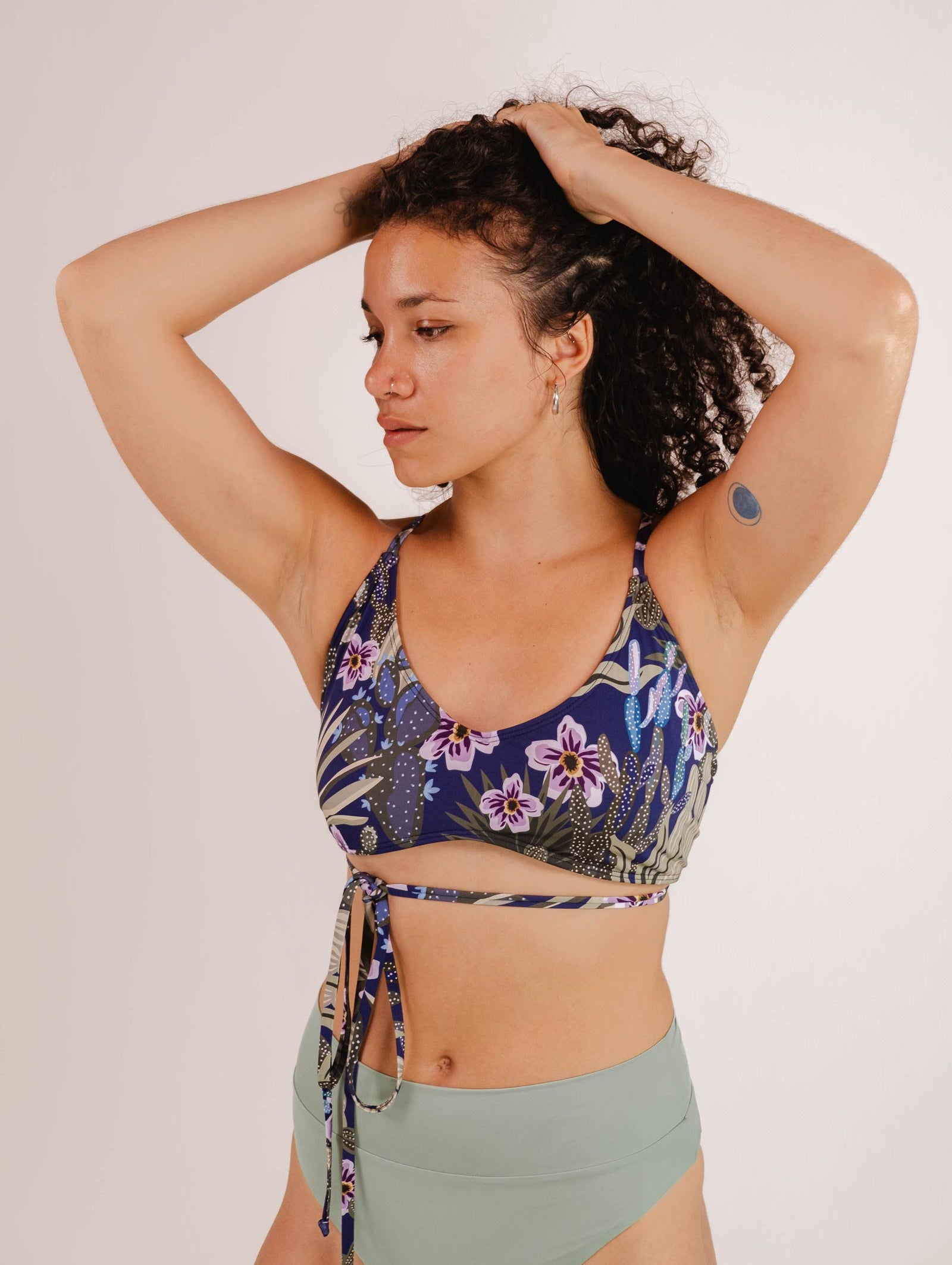 A woman in a Chichi Jardin de Nuit - Bralette Bikini Top by Mimi & August with adjustable straps and green bottoms poses with her hands on her head against a plain background. She has a small bandage on her left arm.