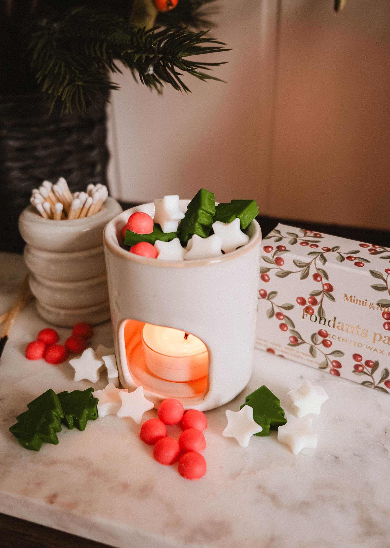 A white ceramic wax melt warmer by Mimi & August glows with a tealight, surrounded by star, tree, and round Christmas Scented Wax Melts in white, green, and red—perfect for adding festive fragrance to your home.