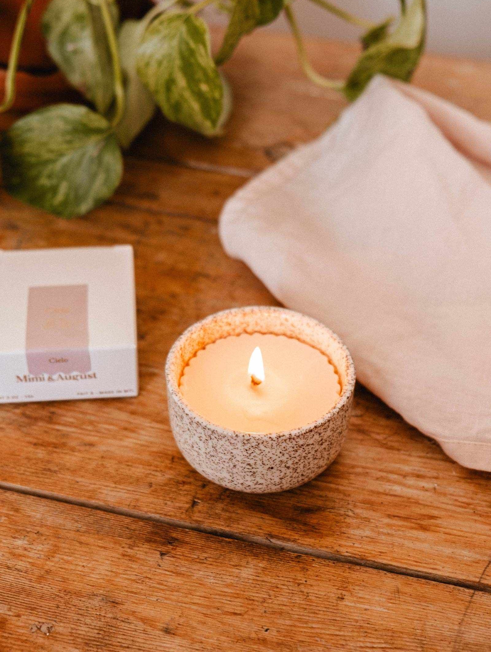 A lit Candle Refill Cielo by Mimi & August glows in a speckled ceramic holder on wood beside a white cloth and a box labeled "Mini August," with green leaves in the background.