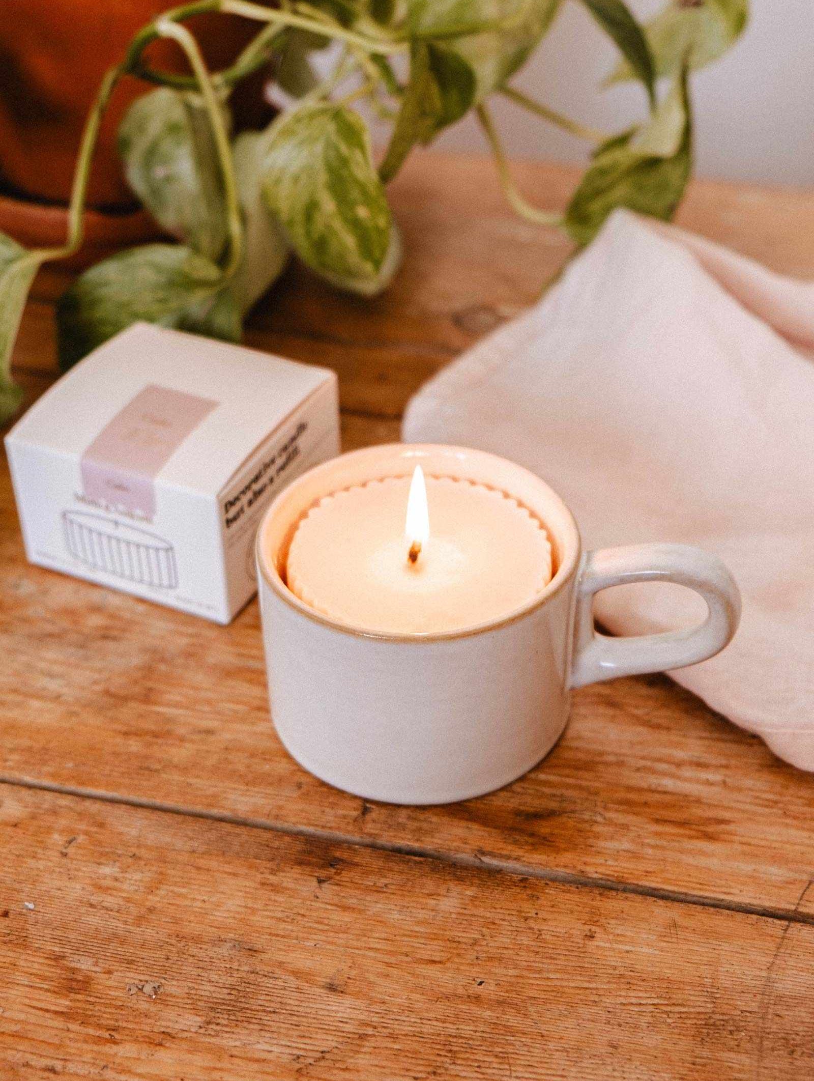 The Candle Refill Cielo by Mimi & August, a premium fragrance soy wax candle refill, rests in a ceramic mug on a wooden table with a white cloth, box, and green potted plant—ideal for refreshing your favorite vessel.