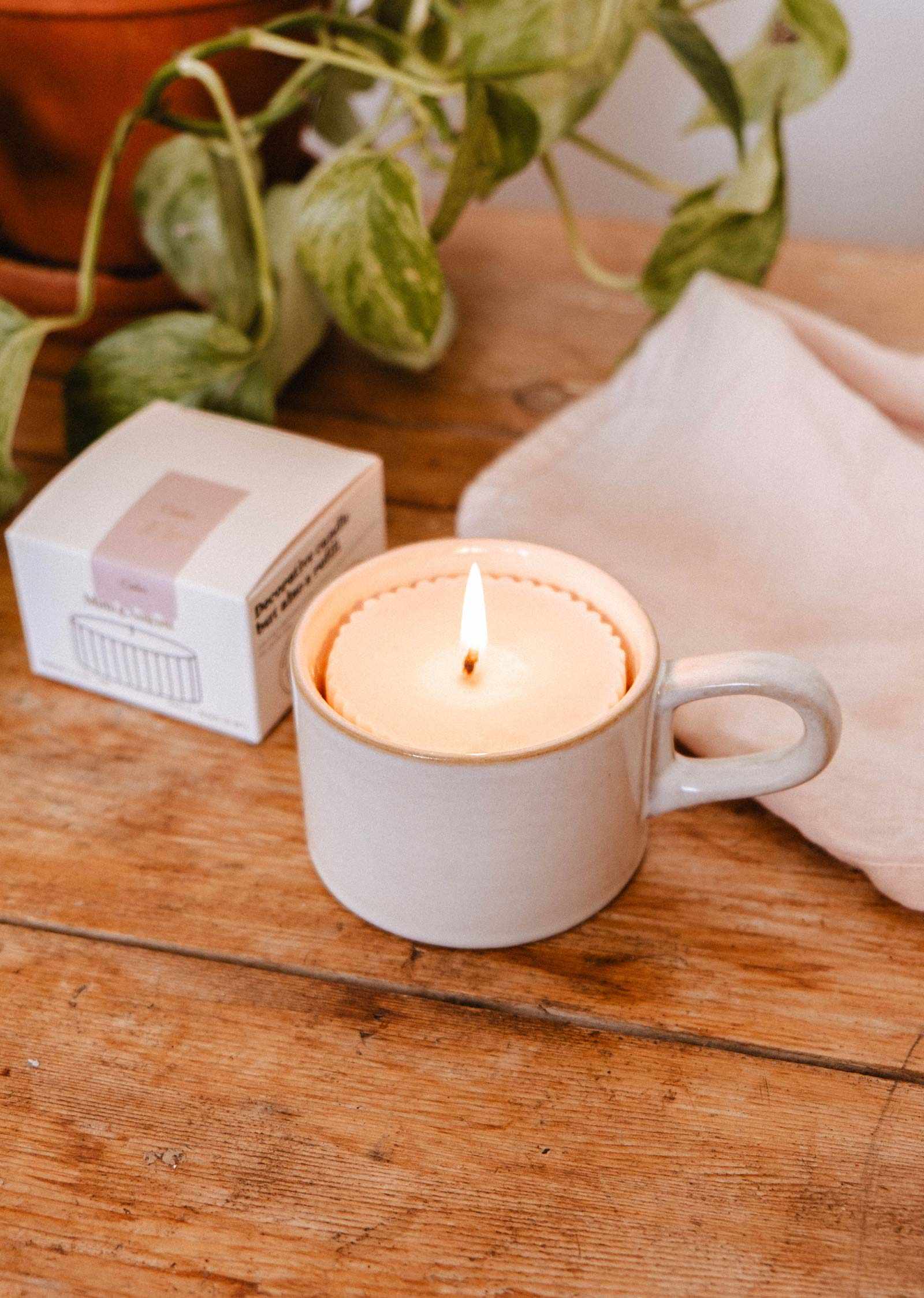 The fig candle refill by Mimi & August, a premium fragrance soy wax candle refill, rests in a ceramic mug on a wooden table with a white cloth, box, and green potted plant—ideal for refreshing your favorite vessel.