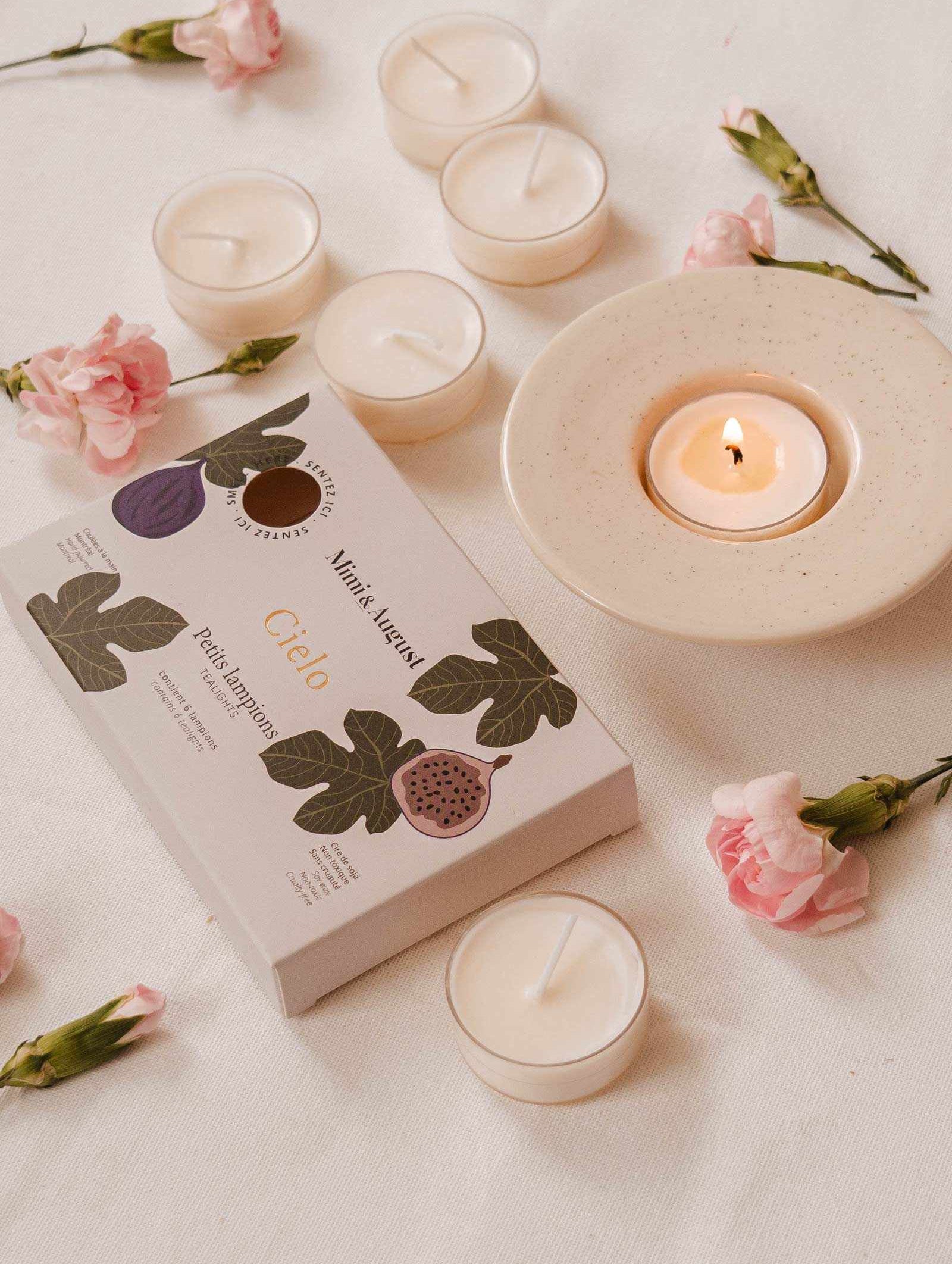 A lit tea light candle on a ceramic holder amidst pink flowers creates a cozy ambiance. The Cielo Tealight Candle Set from Mimi & August, promising a warm comforting scent, sits gracefully on the white surface.