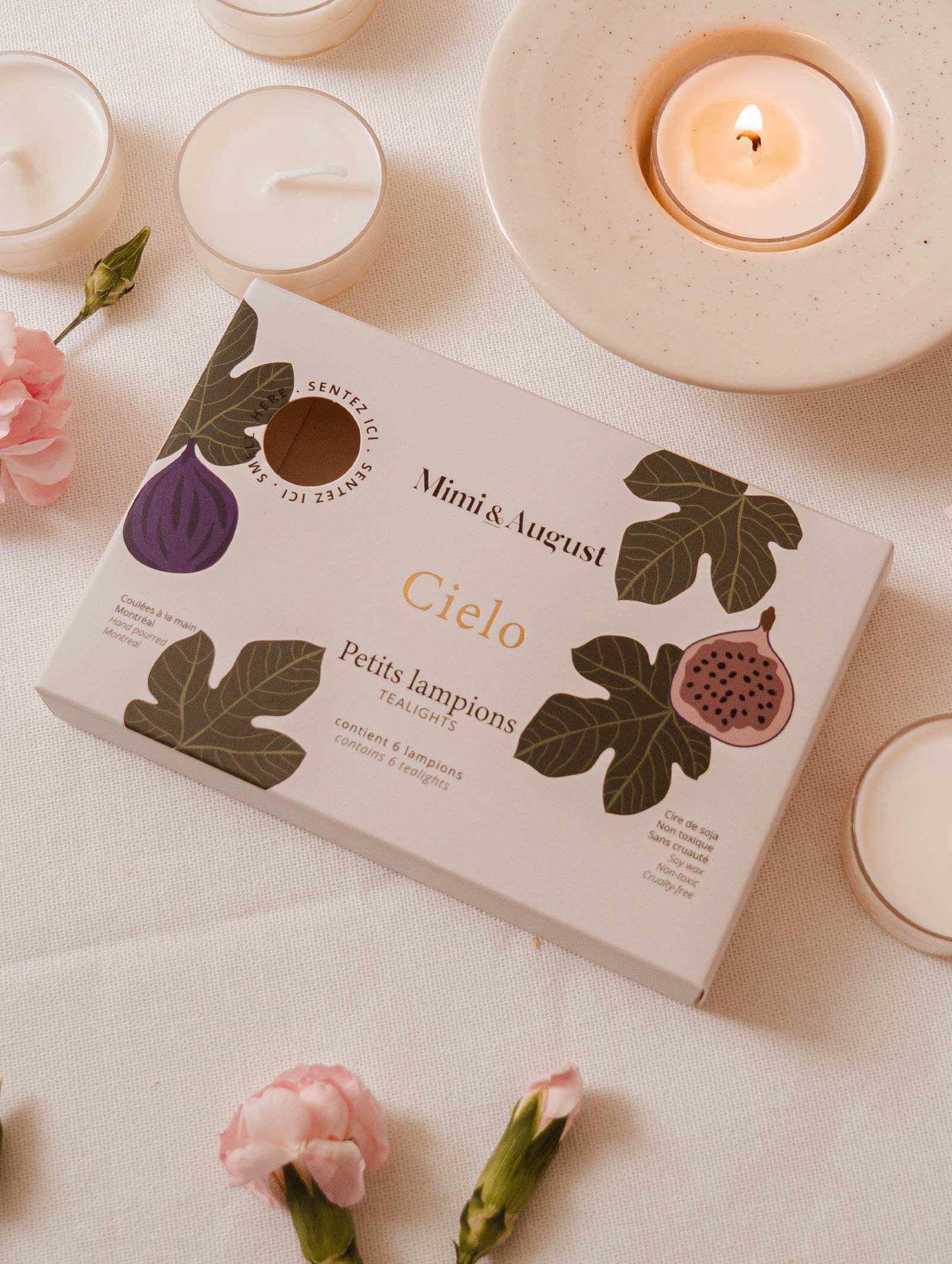The Cielo Tealight Candle Set by Mimi & August, when paired with soft pink flowers and shimmering tea lights, creates a cozy atmosphere. Its warm, comforting scent enhances the inviting ambiance beside a lit candle on a saucer.