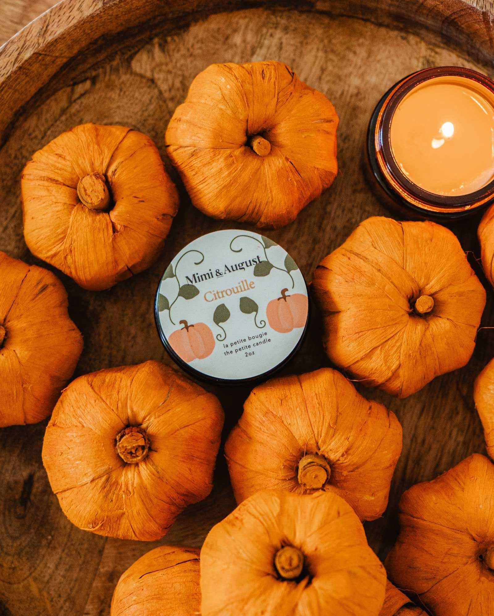 A collection of small orange pumpkins surrounds the Mimi & August Citrouille - Reusable Candle in a wooden bowl, exuding a warm and soothing pumpkin fragrance.